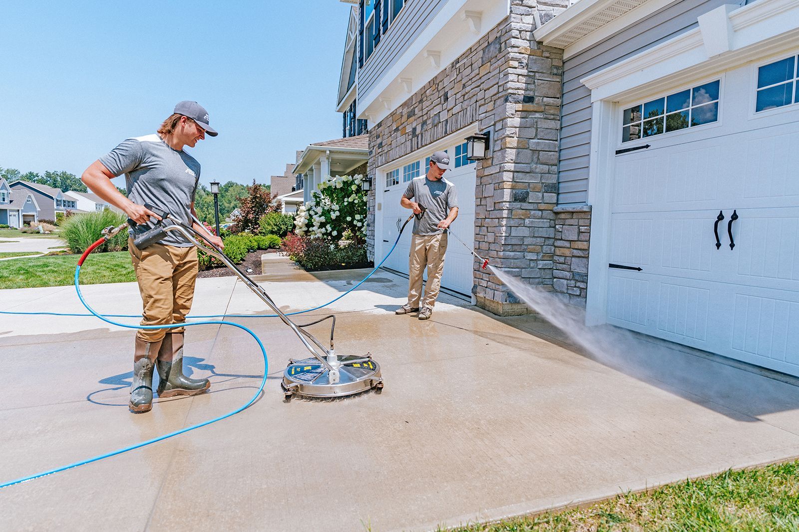 Two service workers use surface cleaners on a residential concrete driveway in front of a house with a stone facade.