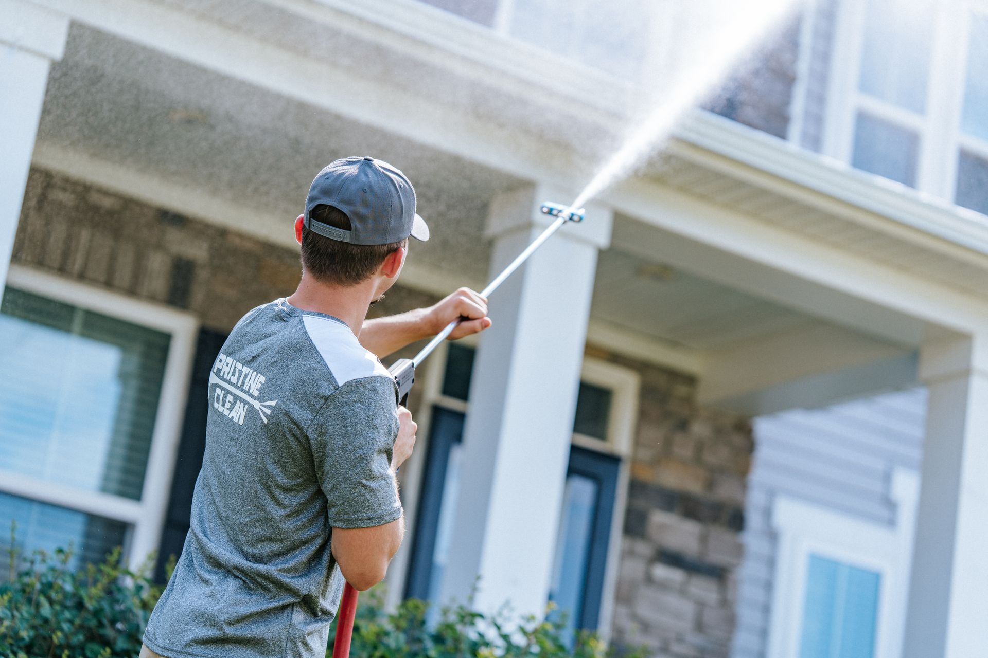 A worker in a gray uniform power washes the exterior of a multi-story house.