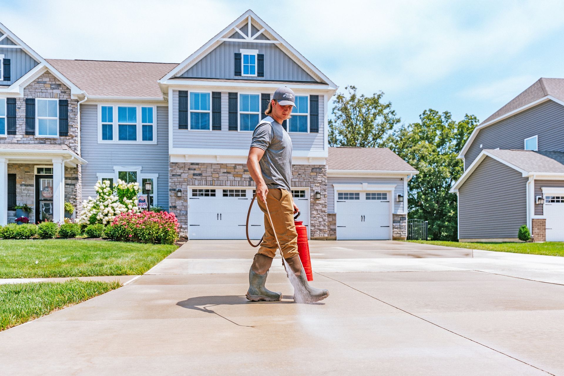 A person in work gear uses a pressure washer to clean a concrete driveway in front of a suburban house.