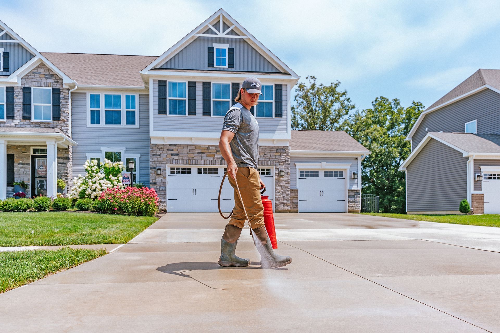 A person in a gray t-shirt and tan pants uses a power washer to clean a concrete driveway in front of a house.
