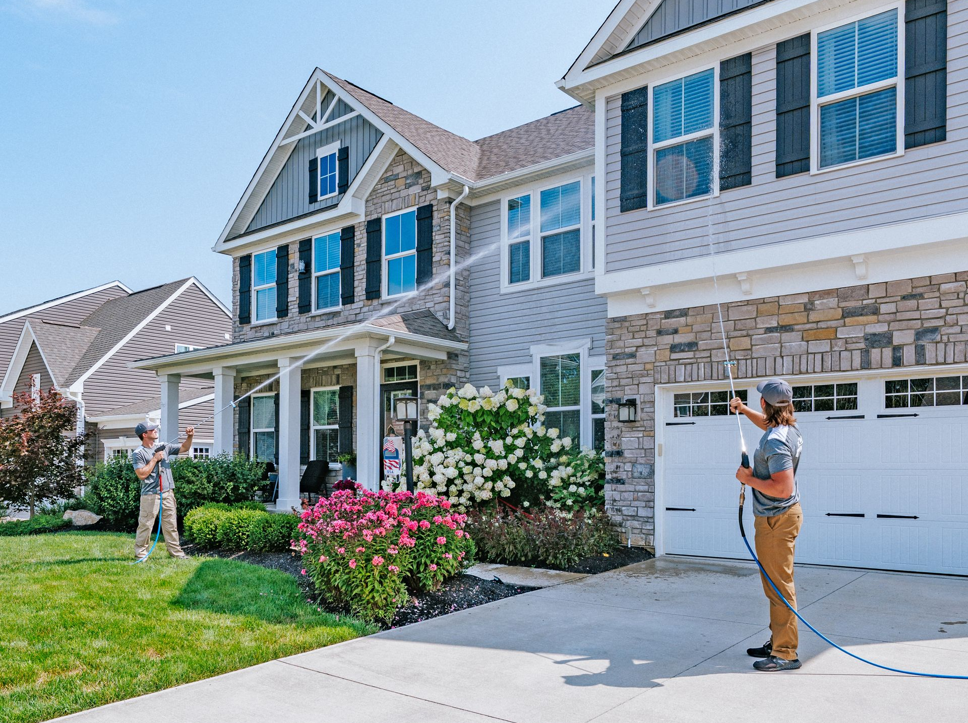 Two people pressure wash the exterior of a suburban, two-story house with siding and stone accents on a sunny day.
