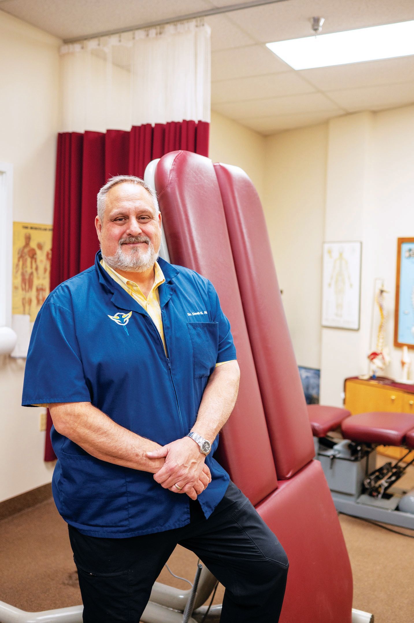 A smiling clinician in a blue scrub top leans against a maroon treatment table in a medical examination room.
