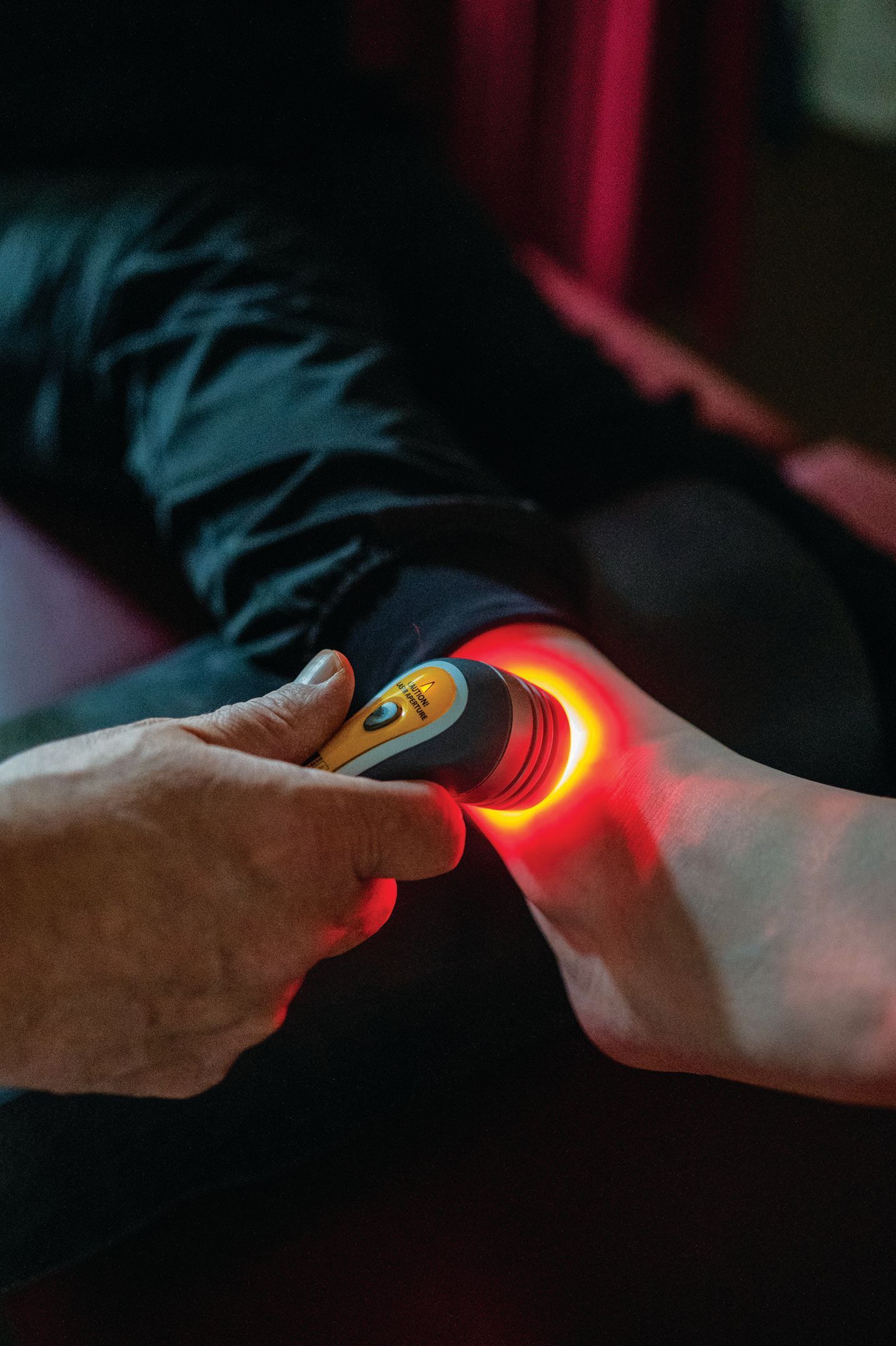 A hand uses a handheld device emitting a bright red light on a person's ankle.