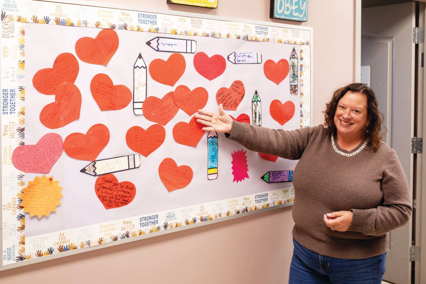 A smiling person gestures toward a classroom bulletin board decorated with numerous red paper hearts and pencil drawings.