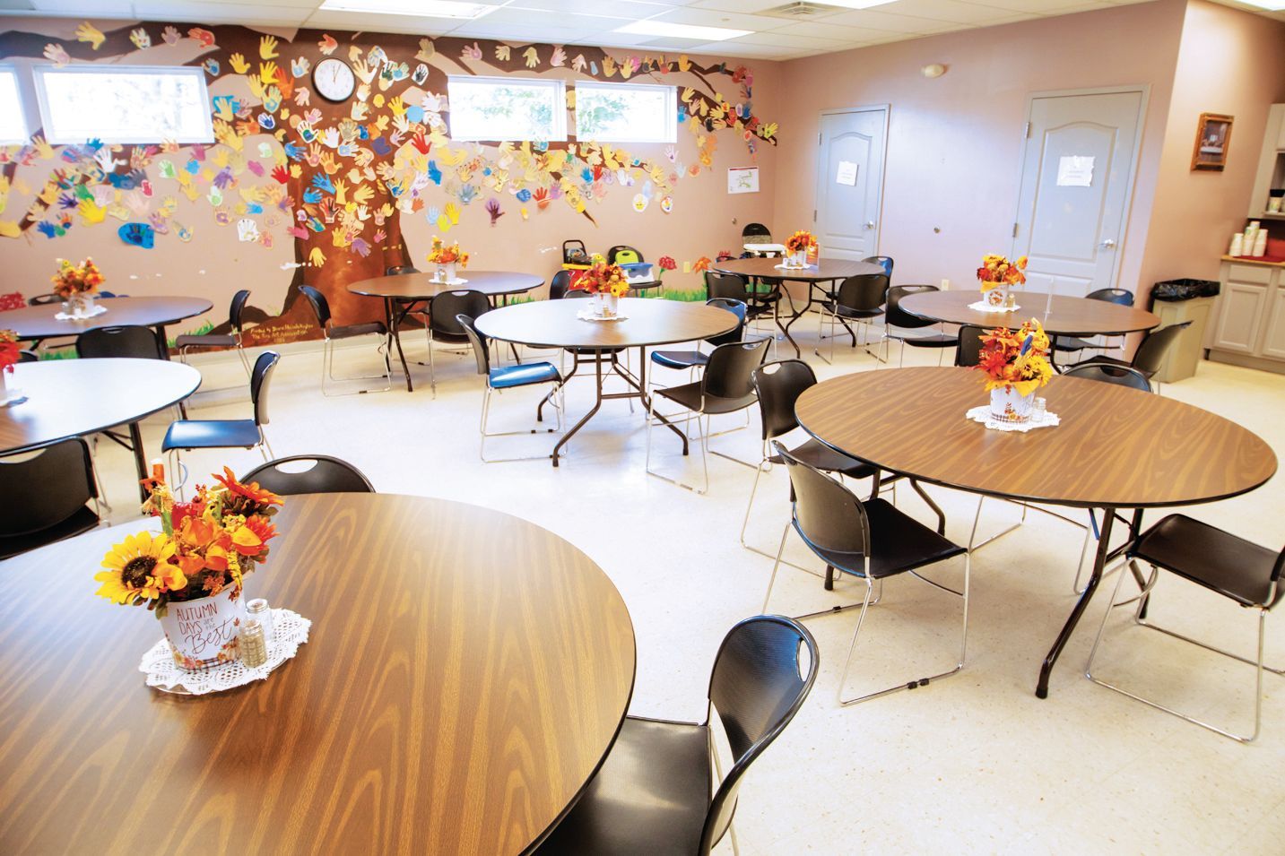 A room with several round wooden tables, black chairs, and autumn-themed floral centerpieces against a mural-painted wall.