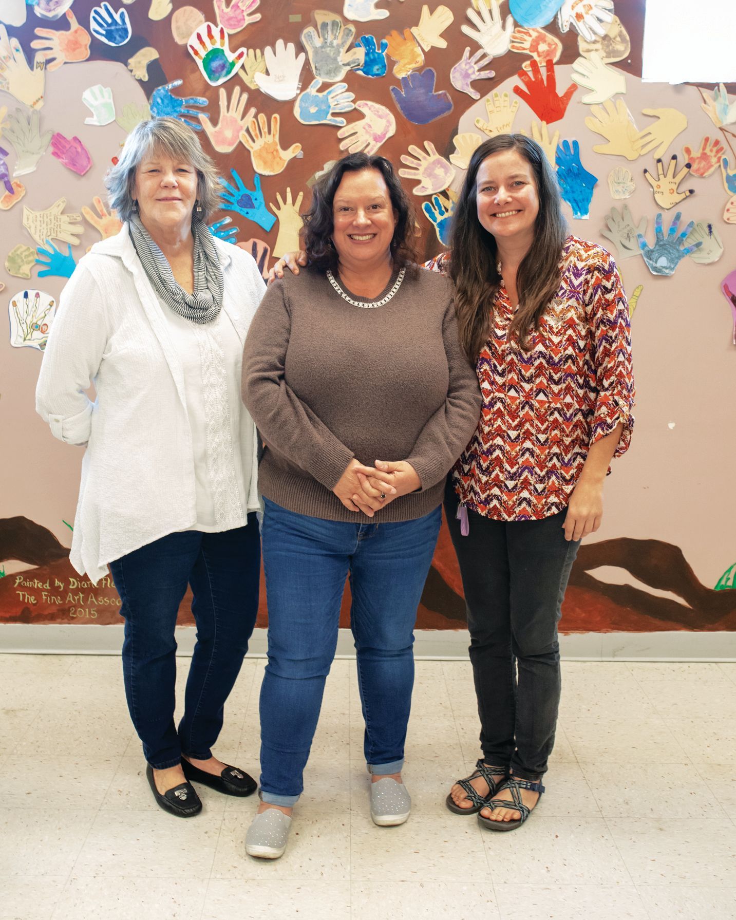 Three people stand side-by-side in front of a wall decorated with colorful paper handprints.