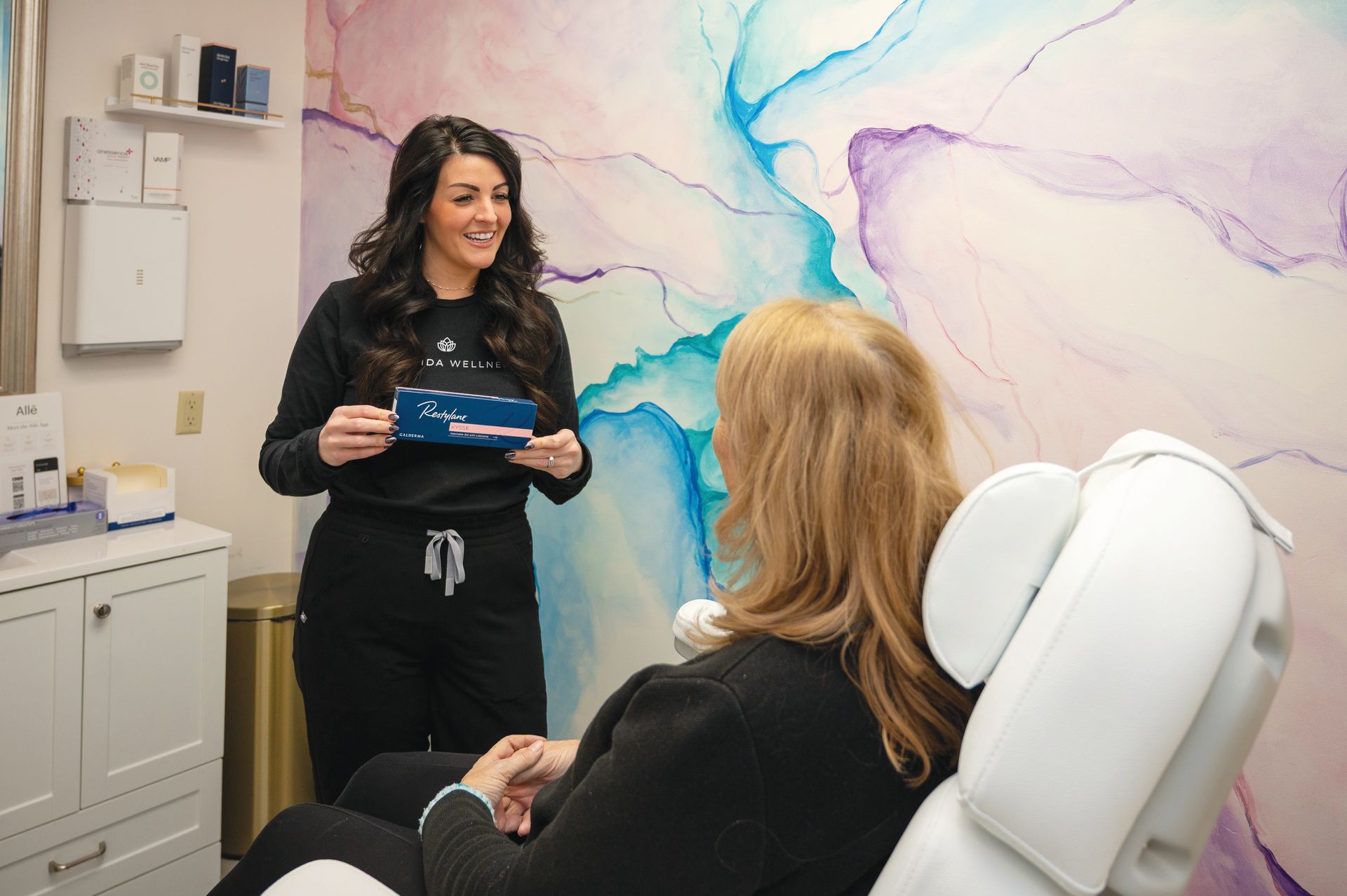 A professional consults with a client in a treatment room, holding a branded product card against a watercolor wall.