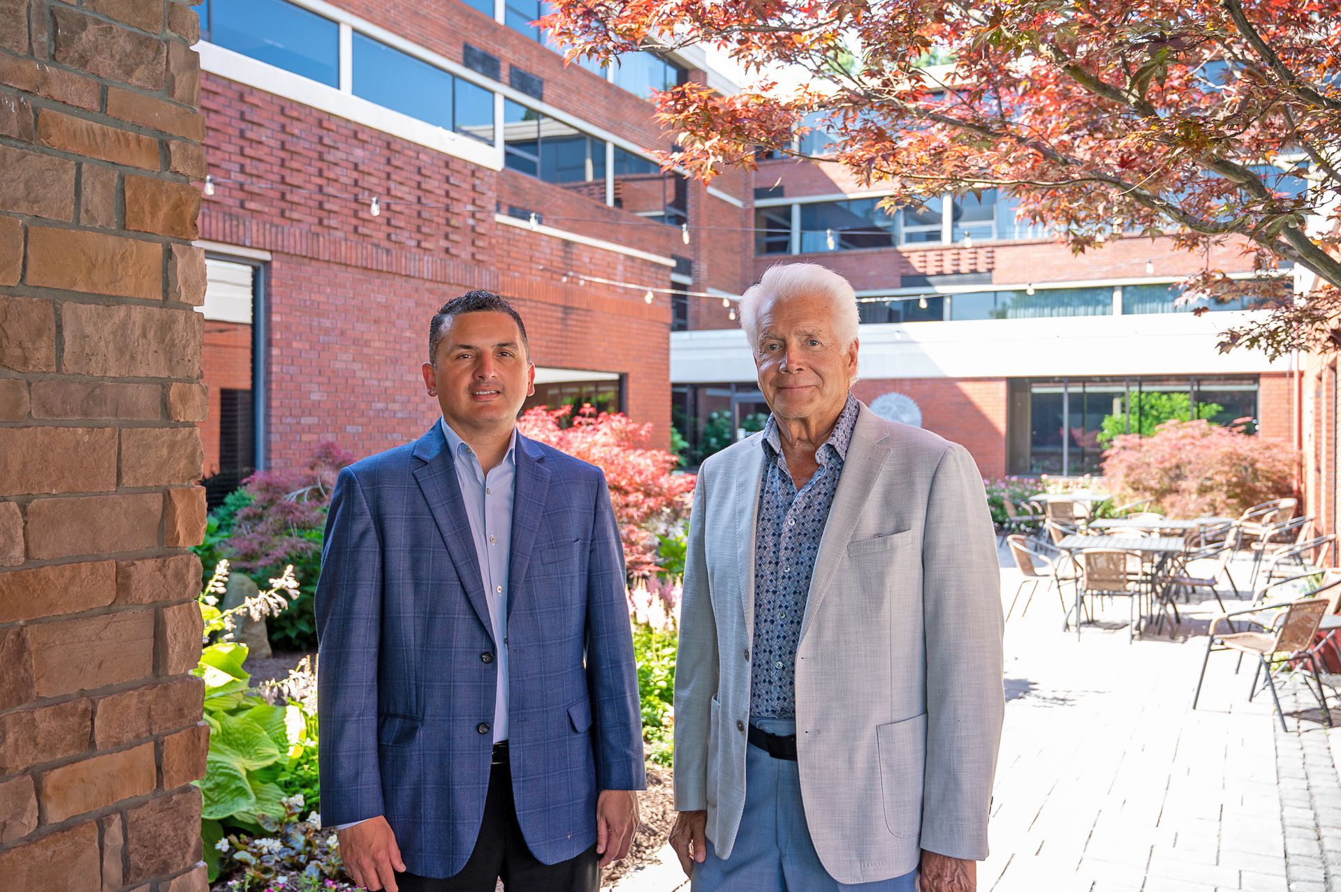 Two men in professional attire stand side-by-side in a sunny, brick-walled courtyard with trees and patio furniture.
