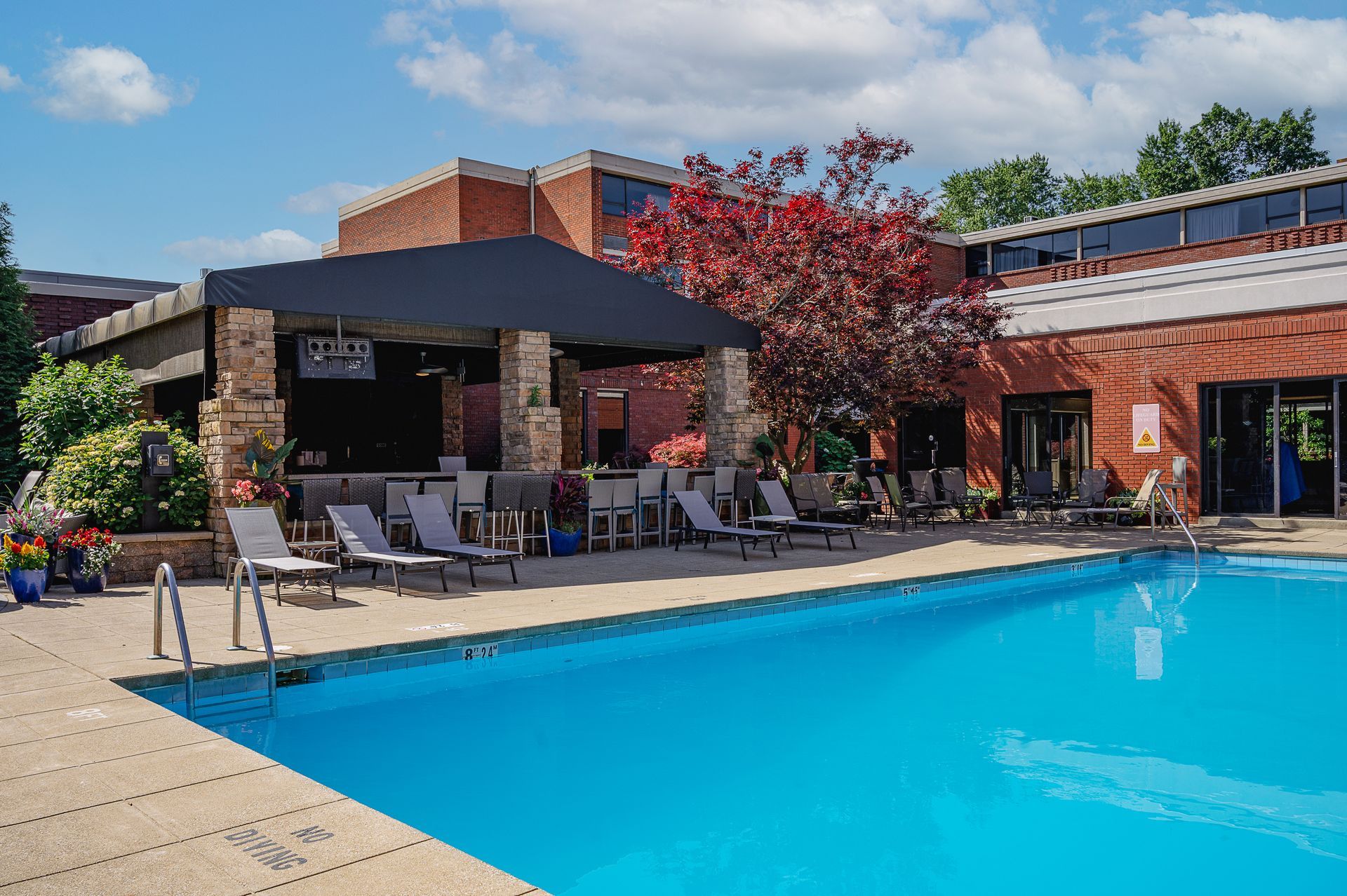 A clear, blue swimming pool sits next to a patio with lounge chairs and a covered outdoor bar area against a brick building.