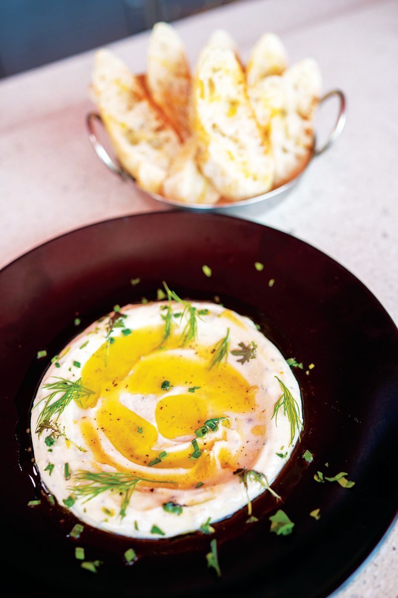 A dark bowl of whipped spread topped with oil and herbs, served with a side of toasted bread in a small metal container.