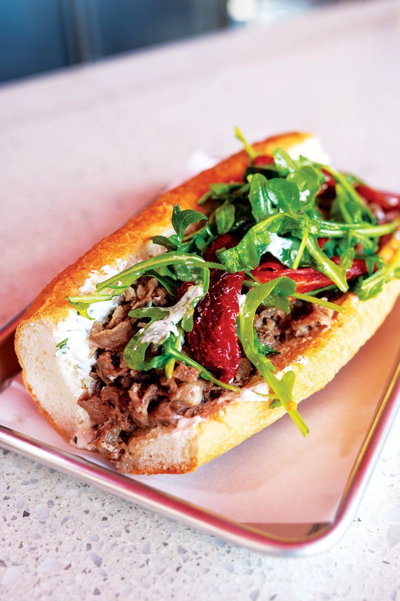 A hoagie-style sandwich topped with arugula and roasted red peppers, served on a metal tray on a speckled surface.