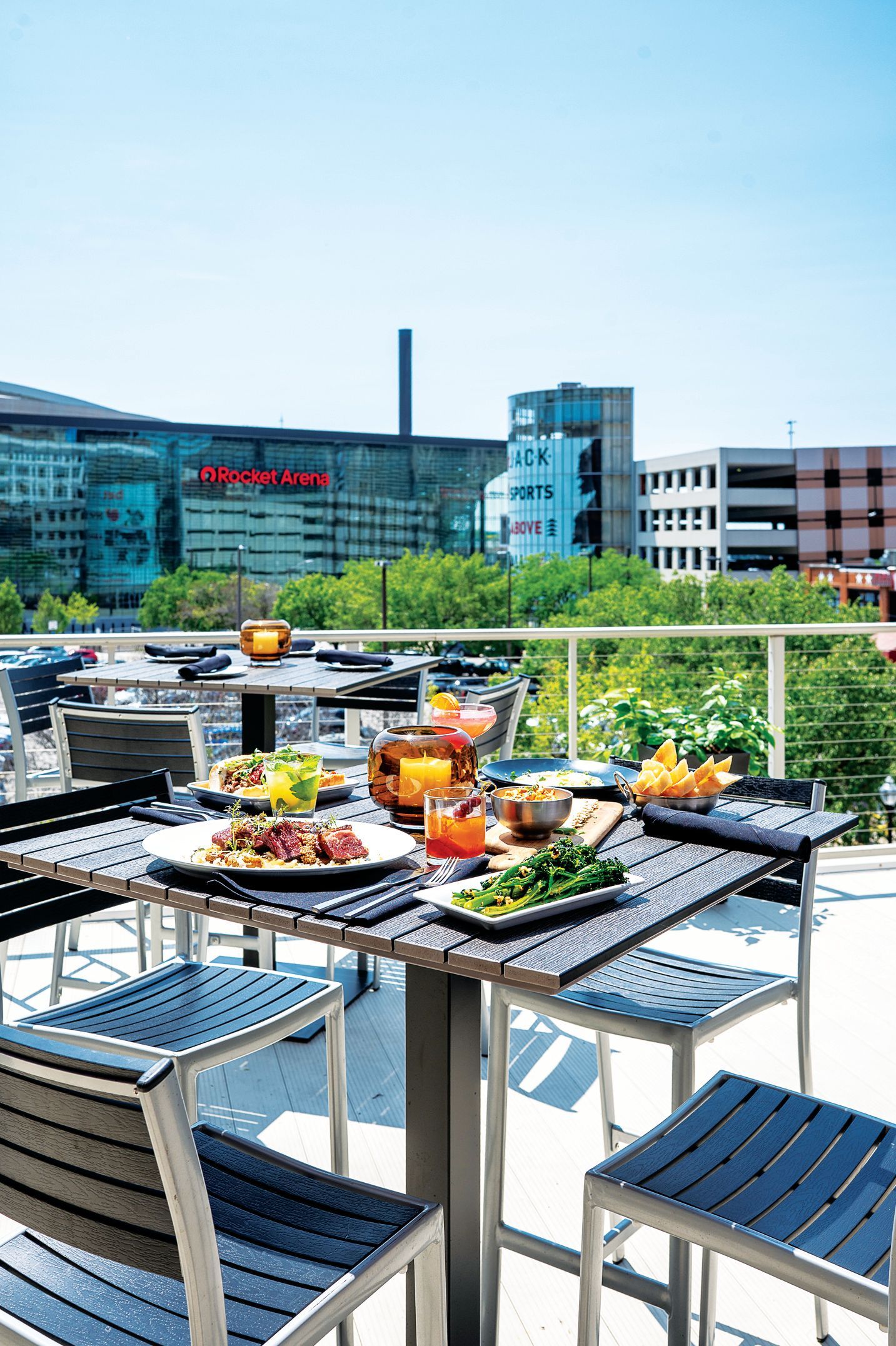 An outdoor rooftop restaurant table with drinks and shared plates, overlooking a city skyline on a sunny day.
