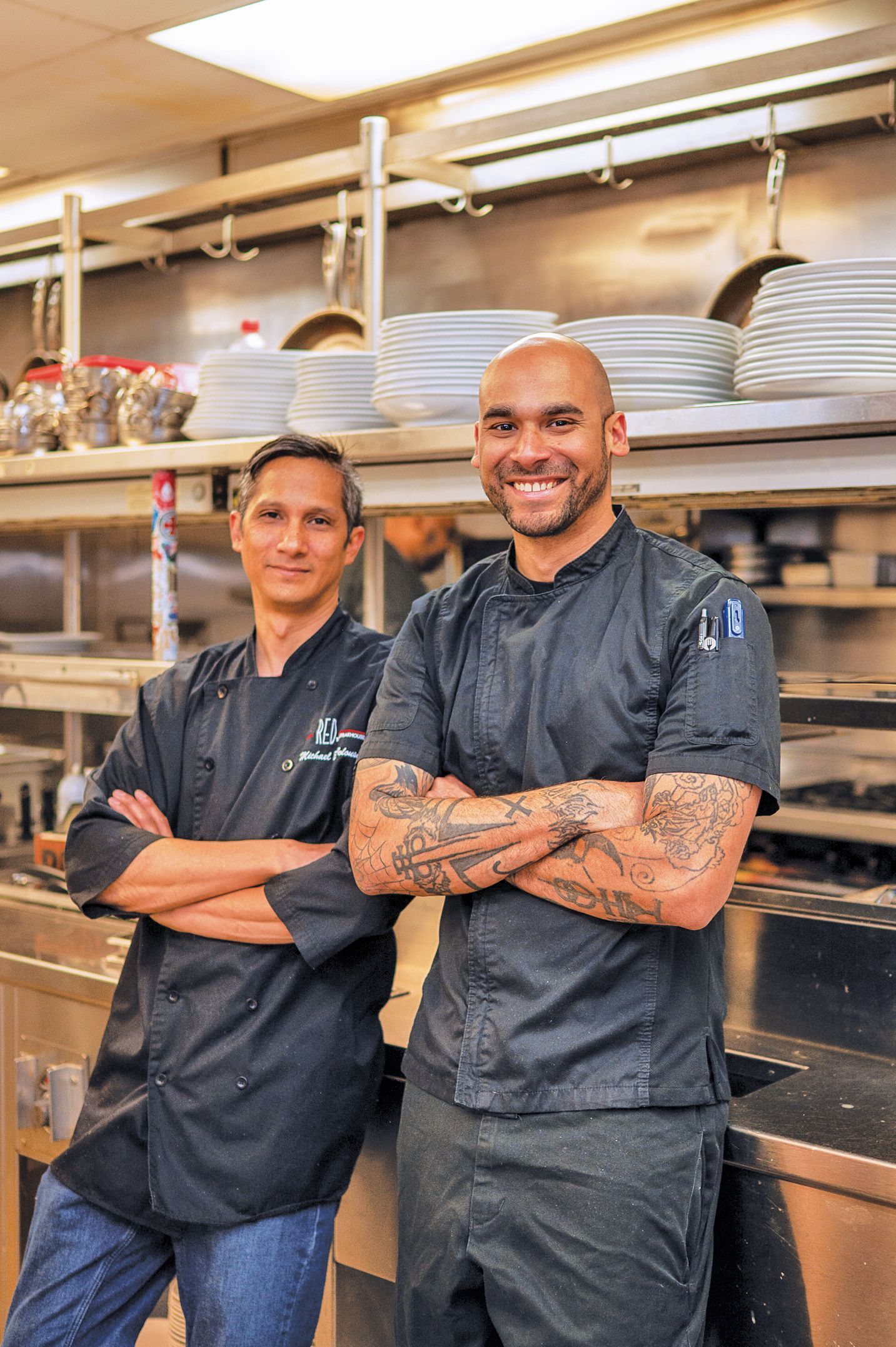 Two smiling chefs in black uniforms and jeans stand with arms crossed in a professional kitchen with stacked plates.
