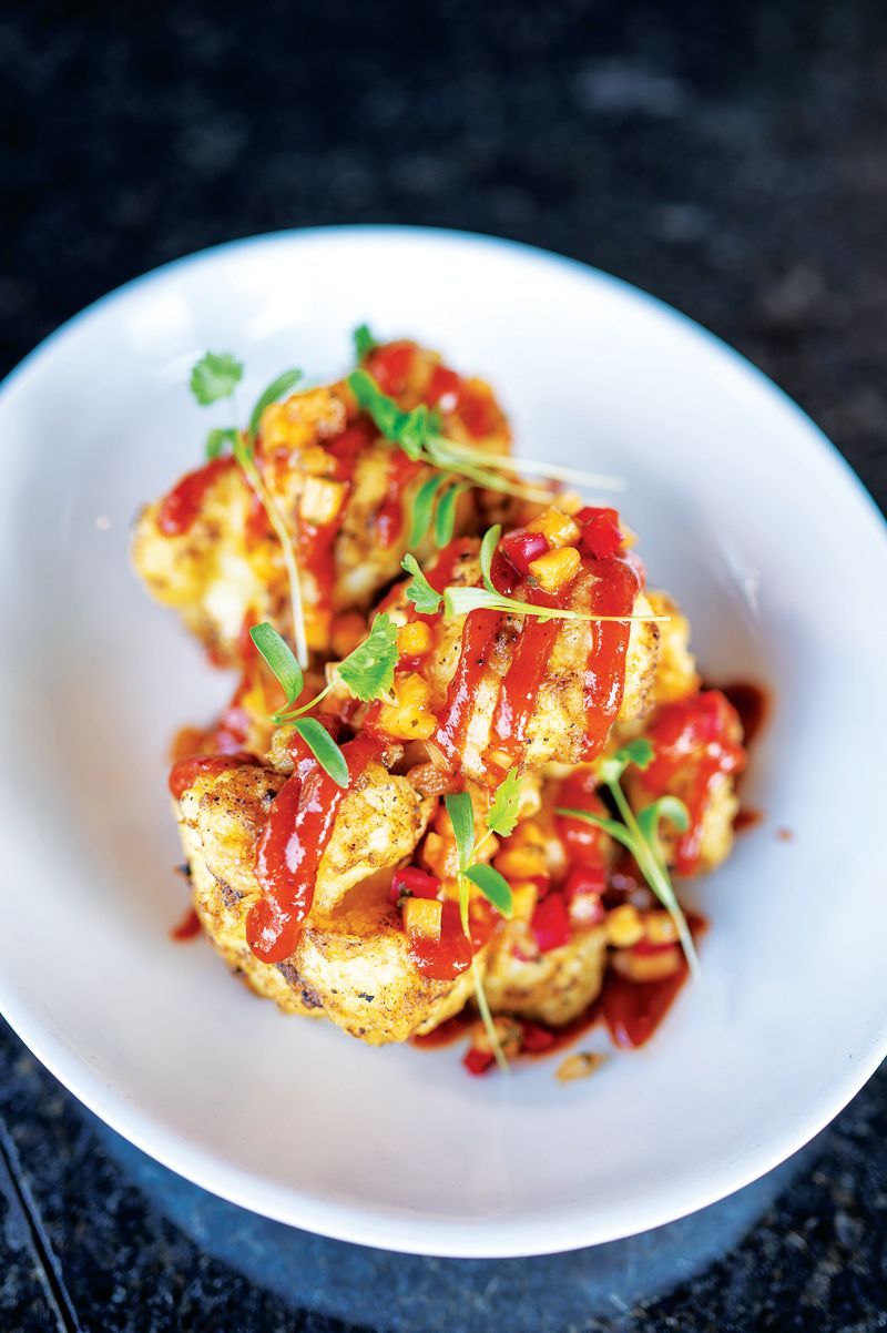 A white plate holds pieces of roasted cauliflower topped with red sauce, yellow diced vegetables, and fresh green herbs.