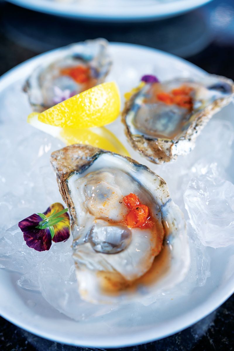 Three raw oysters served on crushed ice with lemon wedges and small purple flowers on a white plate.