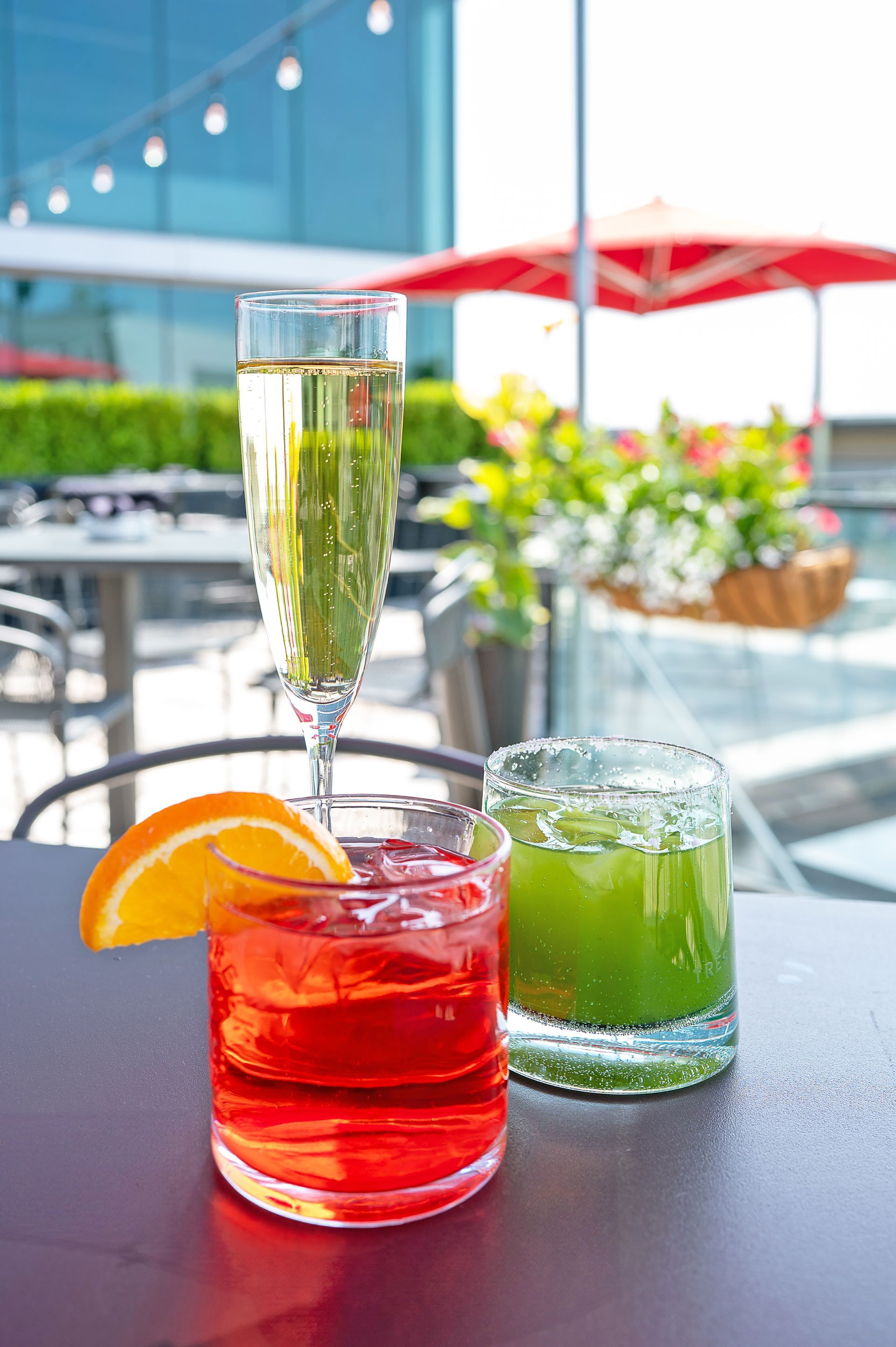 Three cocktails—one champagne flute, one orange drink, and one green drink—on an outdoor patio table under a red umbrella.