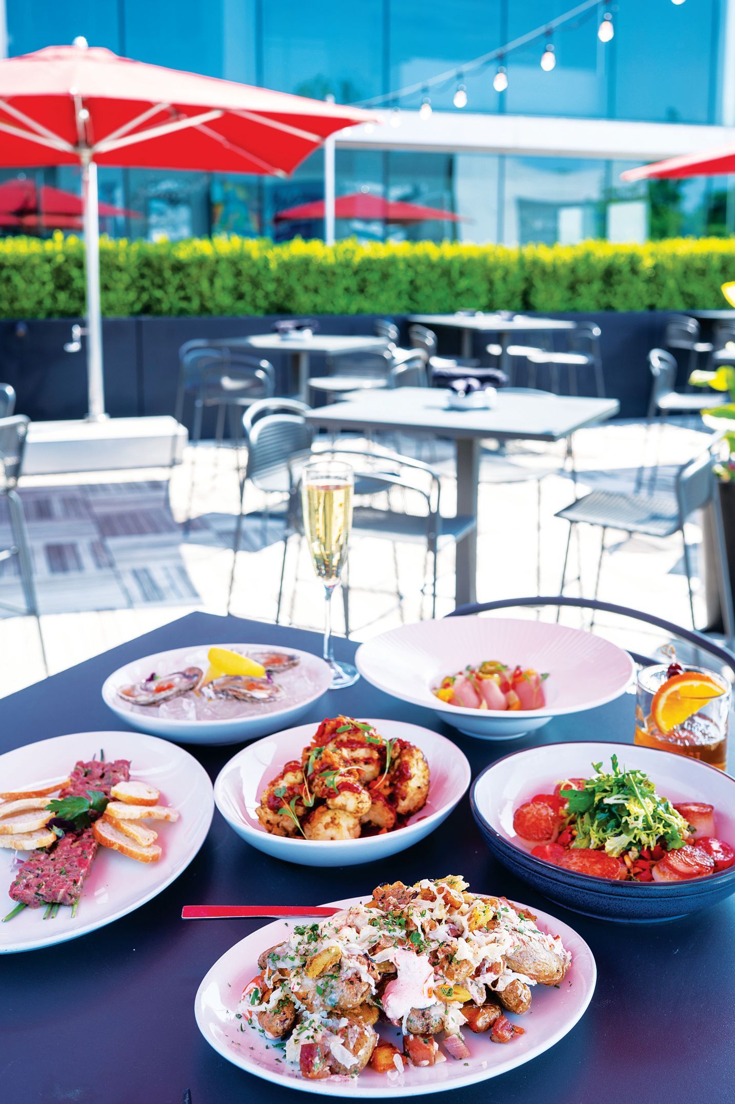 A table on an outdoor restaurant patio features several plates of appetizers, a glass of champagne, and a cocktail.