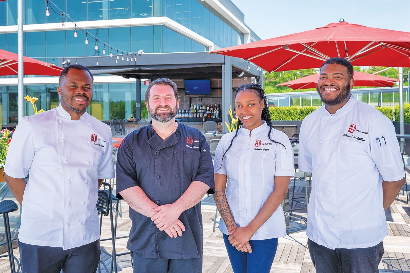 Four chefs in uniforms stand smiling in an outdoor patio setting with red umbrellas and a modern glass building behind them.