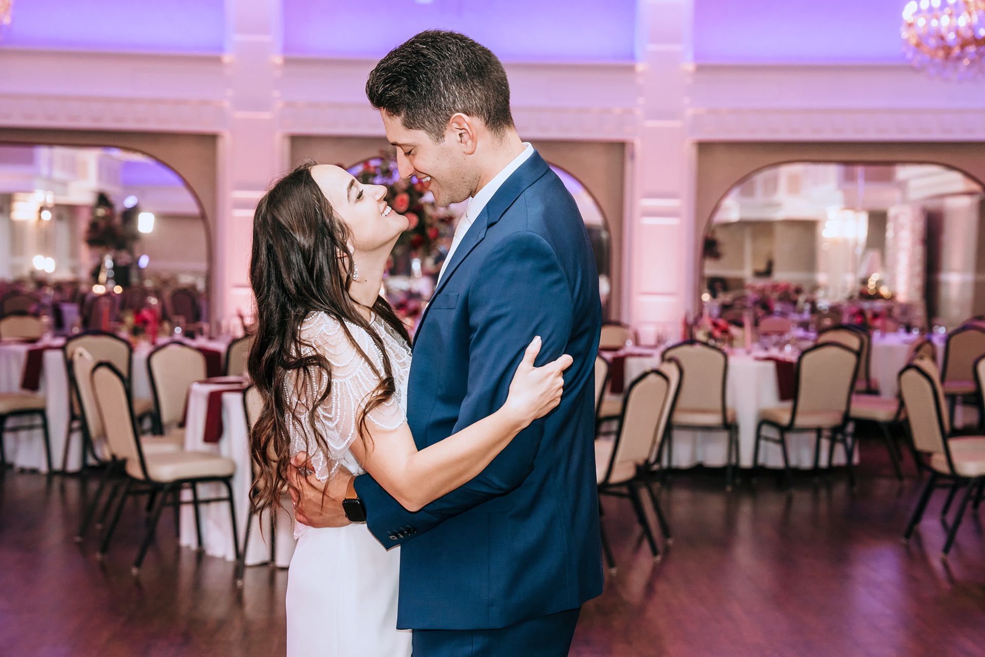 A couple in formal attire embraces and dances together in a brightly lit banquet hall filled with tables and chairs.
