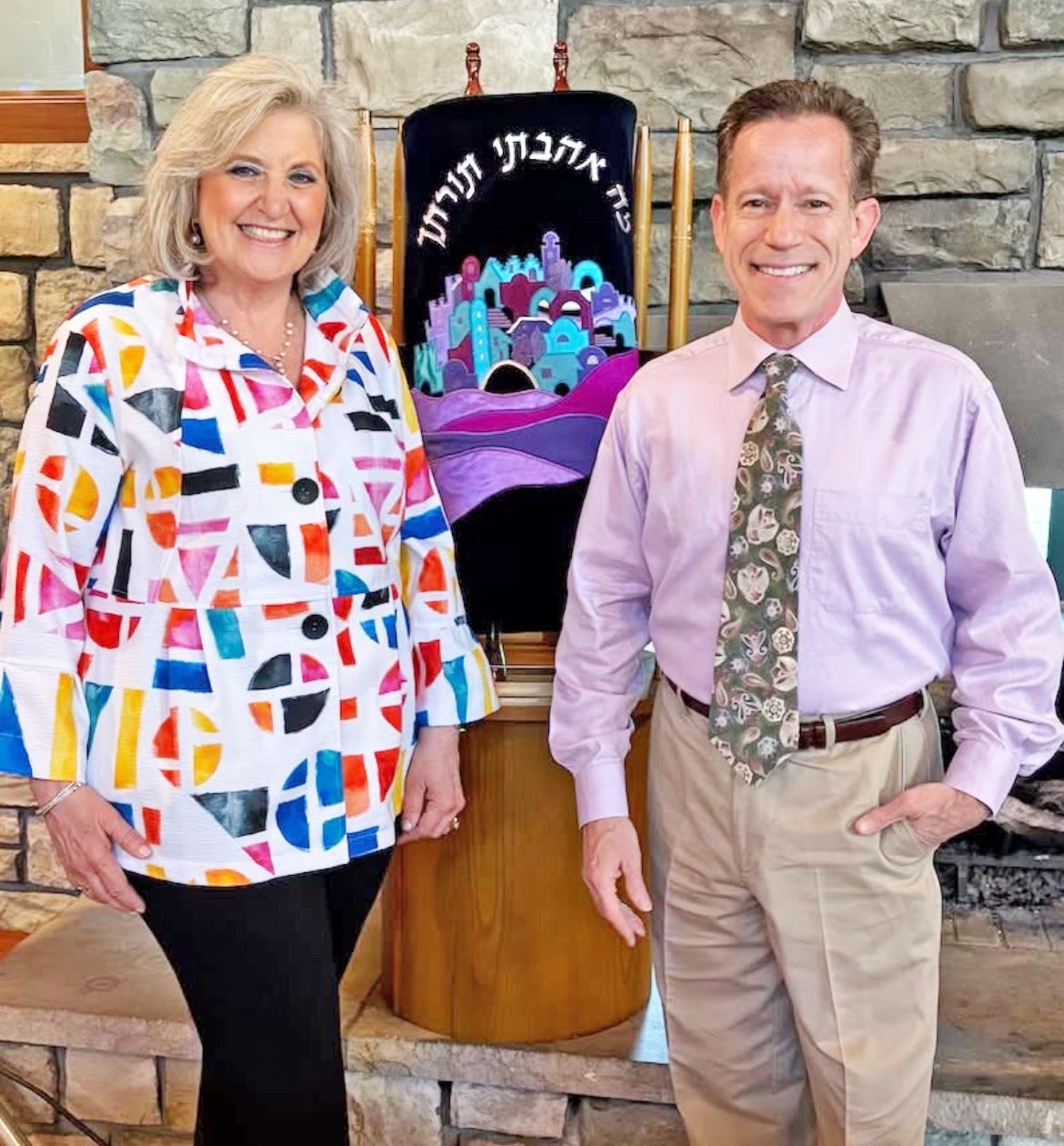 A smiling woman and man stand in front of a colorful Torah mantle with Hebrew text, set against a stone wall backdrop.