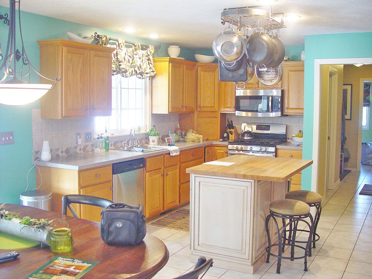 A kitchen with light wood cabinets, teal walls, a center island with bar stools, and a hanging pot rack overhead.