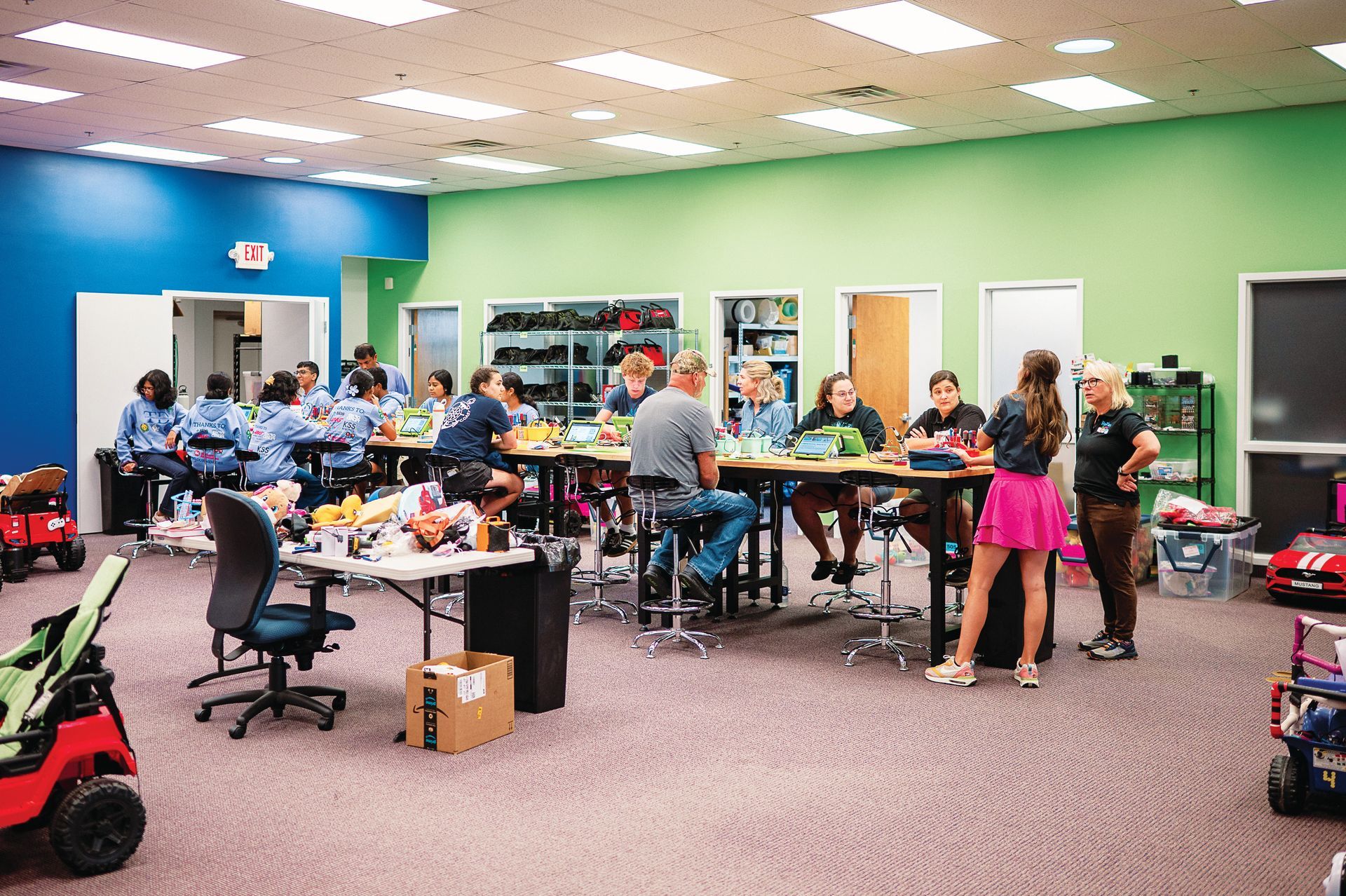 People collaborate in a spacious, colorful room with bright blue and green walls, working at tables with laptops and gear.