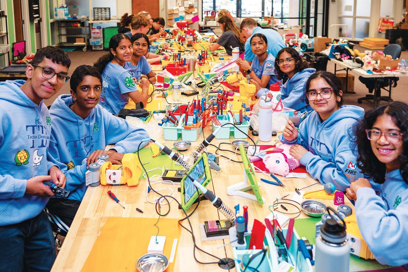 Students in blue hoodies work together at a long table with electronics and tools in a brightly lit classroom.