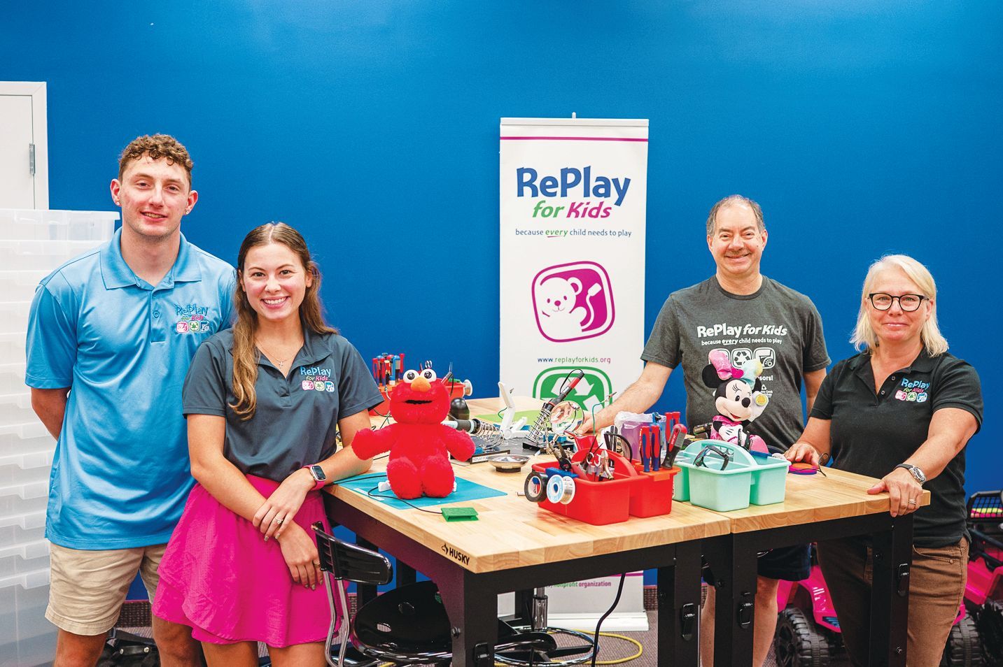 Four people stand behind a wooden workbench with toys and a RePlay for Kids banner against a blue wall.