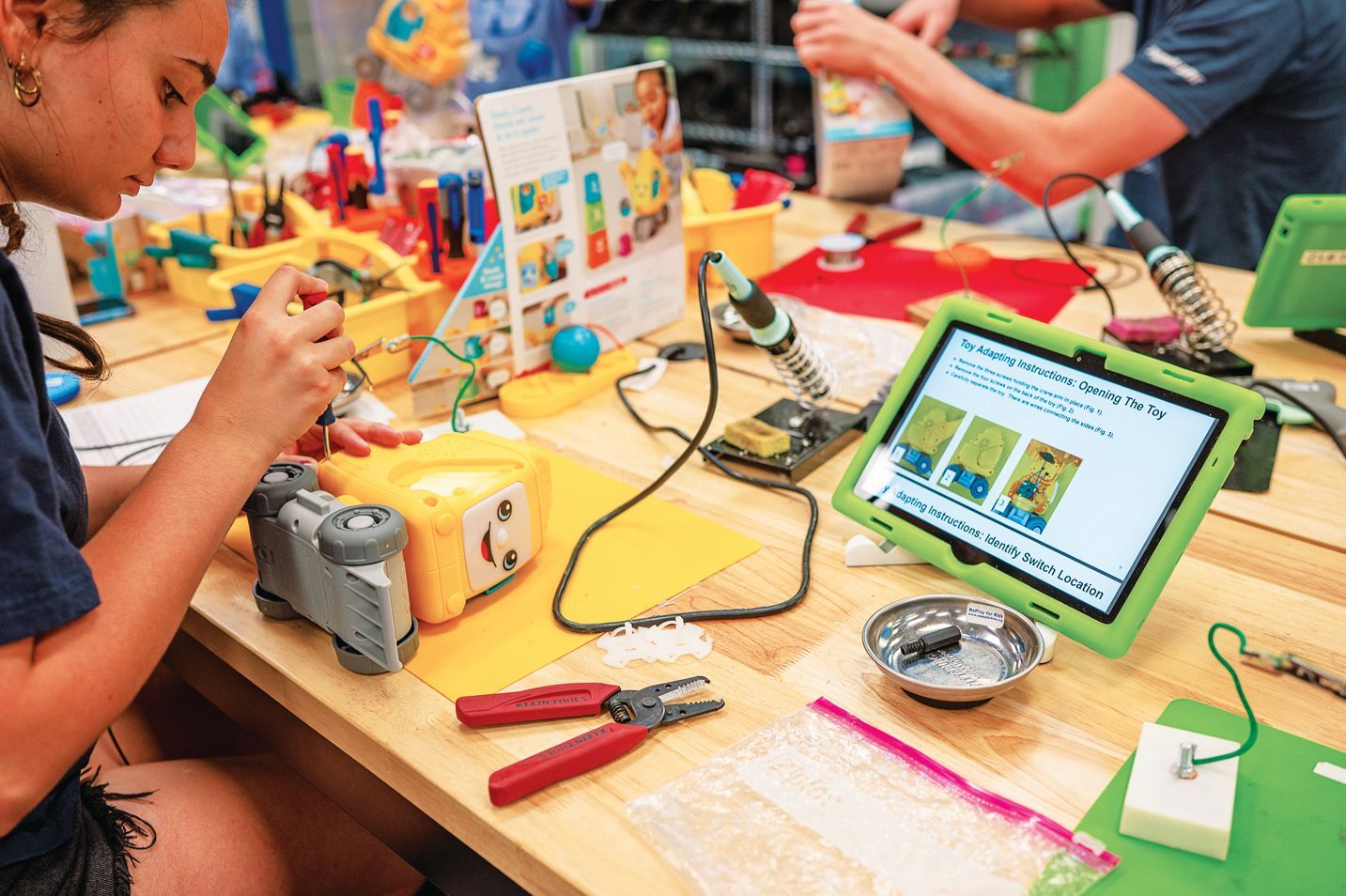 A person uses tools to repair a yellow toy on a wooden table next to a tablet showing instructions and soldering gear.