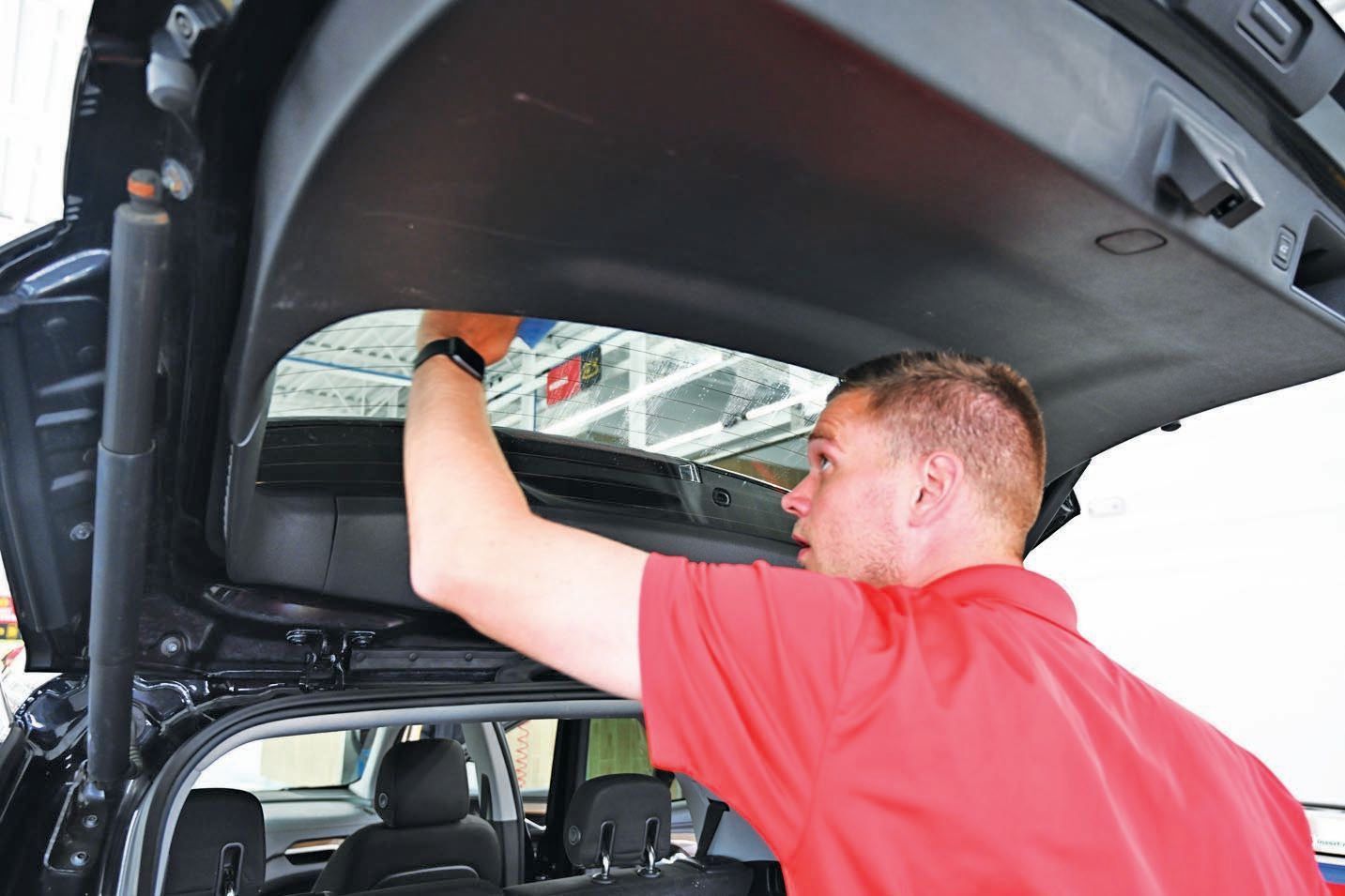 A person in a red polo shirt uses a blue tool to apply tint or a decal to the inside of a car's rear liftgate window.
