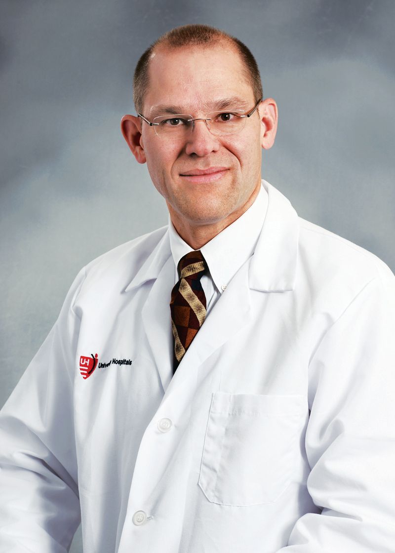 A medical professional in a white coat and tie, smiling against a neutral gray backdrop.