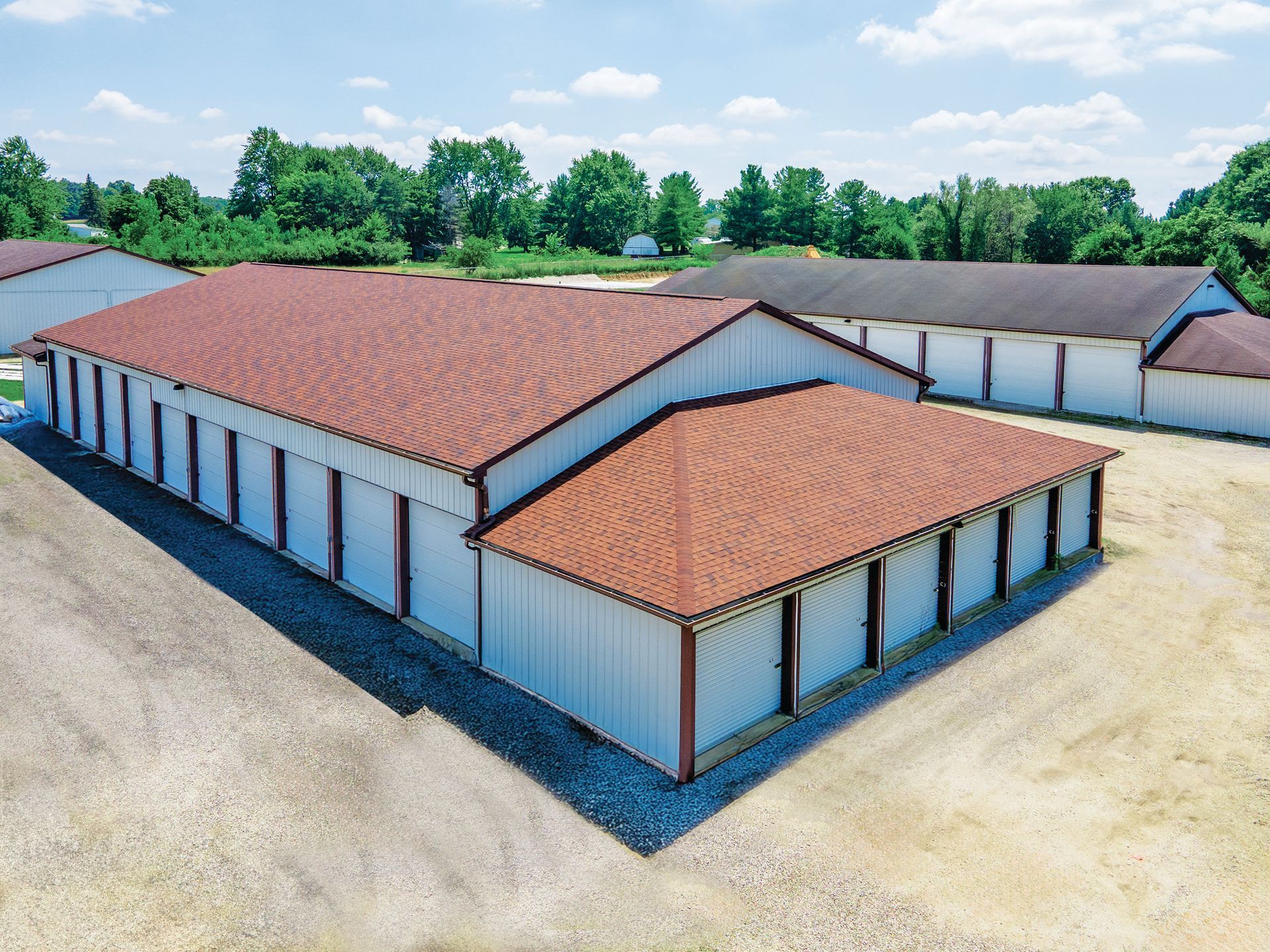 An aerial view of a self-storage facility with white walls and red-brown shingled roofs on a gravel lot under a blue sky.