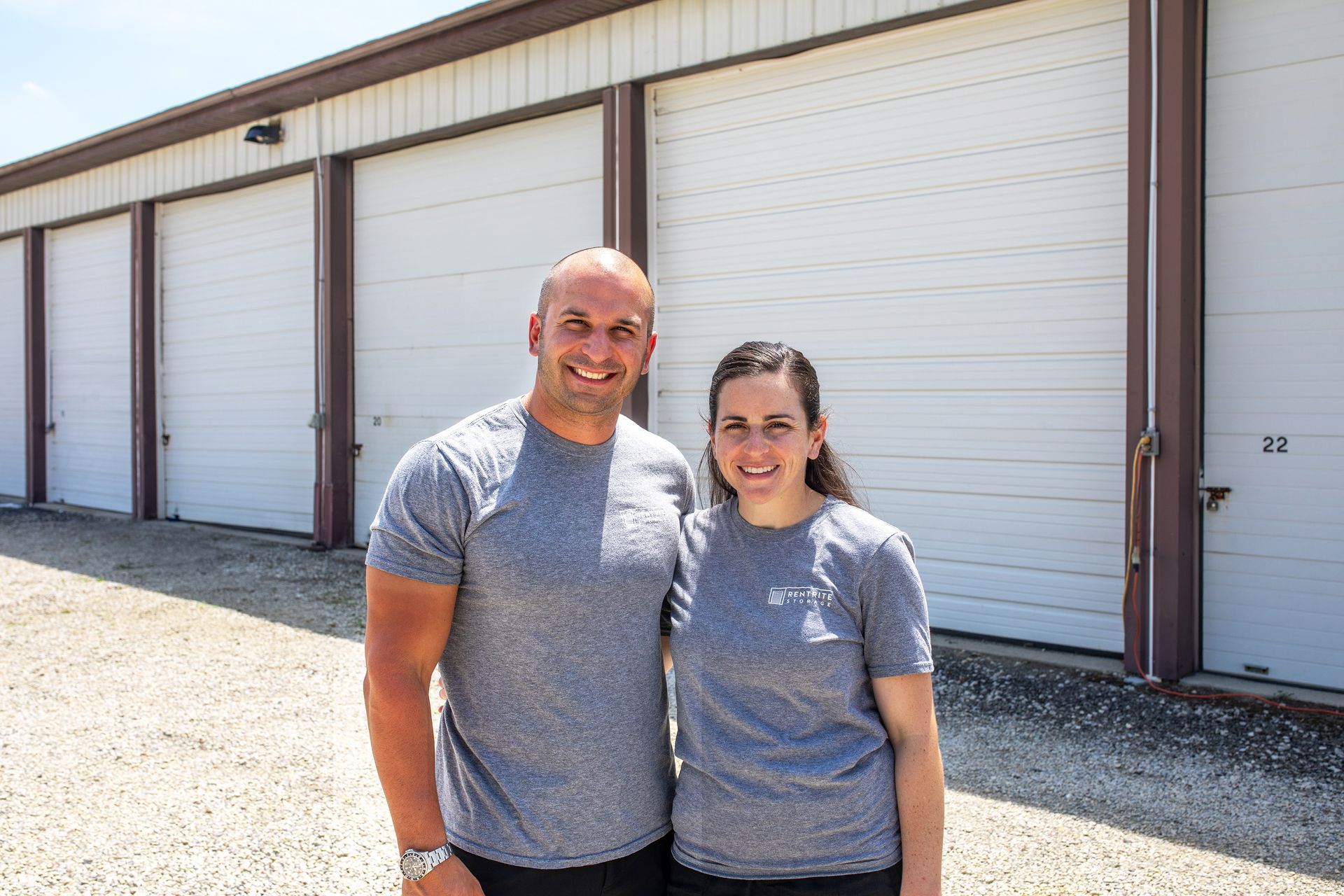 A man and a woman stand smiling in front of a row of white metal storage unit doors at an outdoor facility.