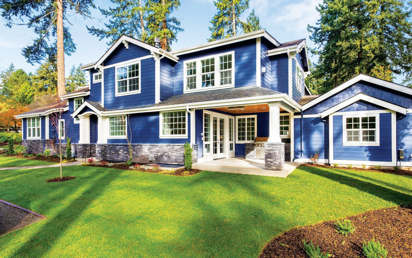 A two-story blue house with white trim and stone accents, set against a green lawn and trees under a sunny sky.