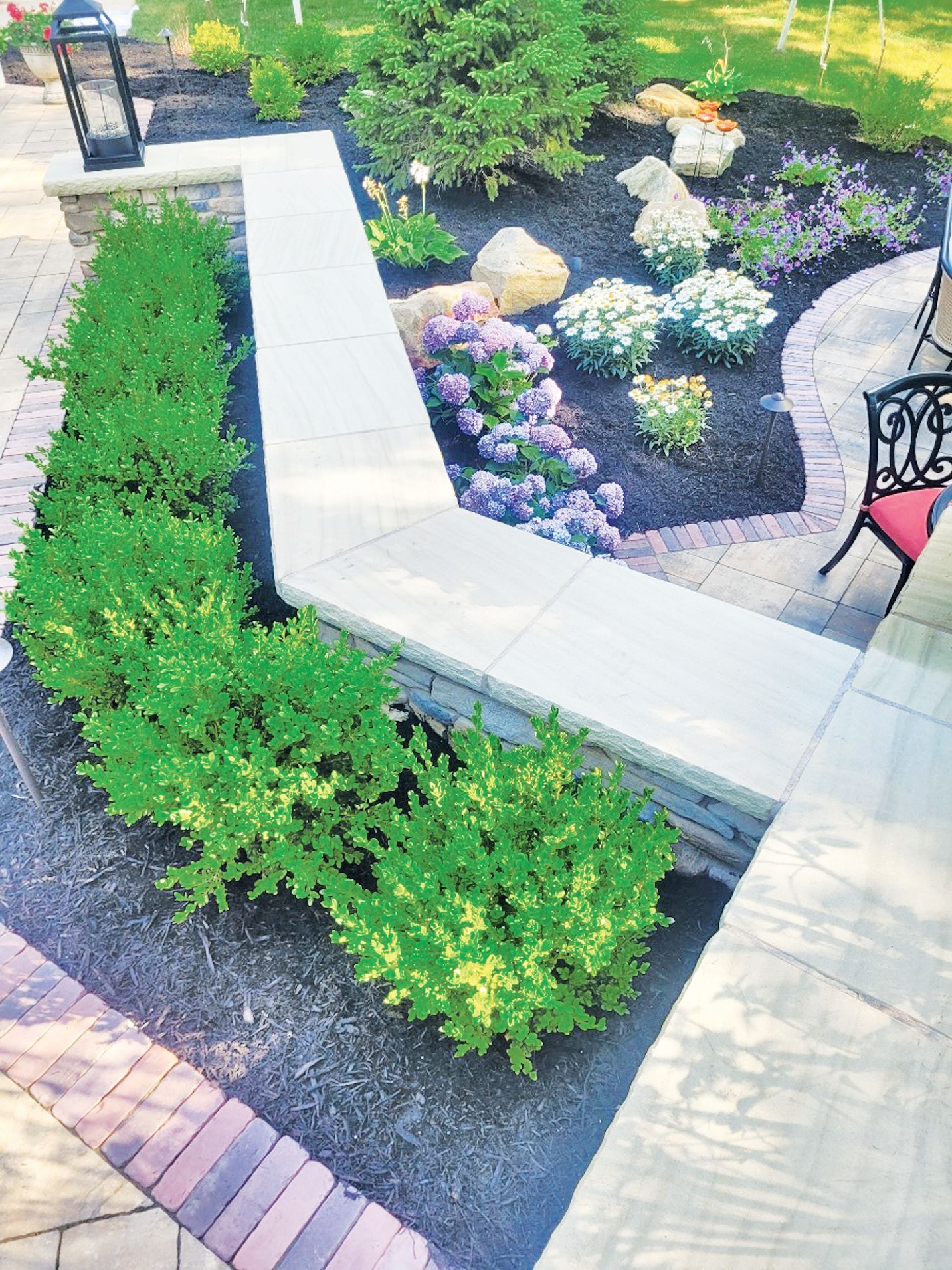 A stone garden wall separates a row of green shrubs from a landscaped garden bed with flowers and white rocks.
