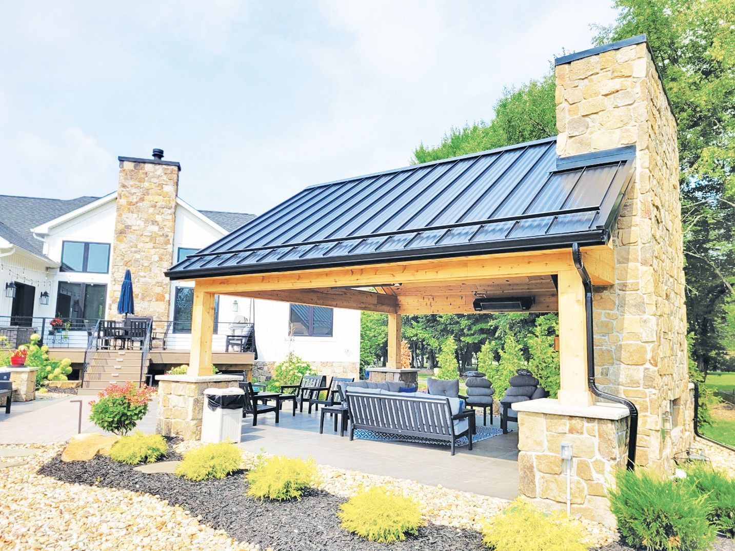 An outdoor covered patio with a stone fireplace, dark metal roof, and patio furniture, next to a house.