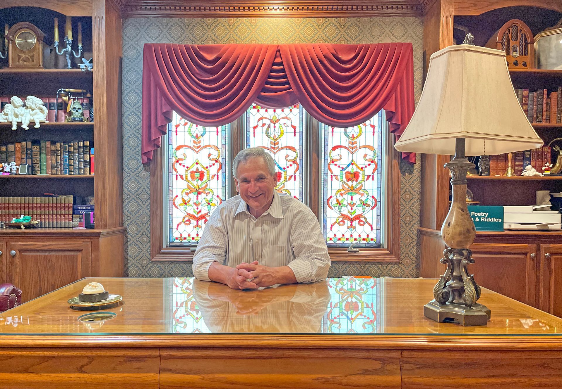 A person sits at a wooden desk in a home office with built-in bookshelves, a stained-glass window, and a table lamp.
