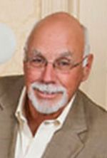 A person with a white beard and glasses wearing a beige jacket and white collared shirt, smiling against a neutral wall.