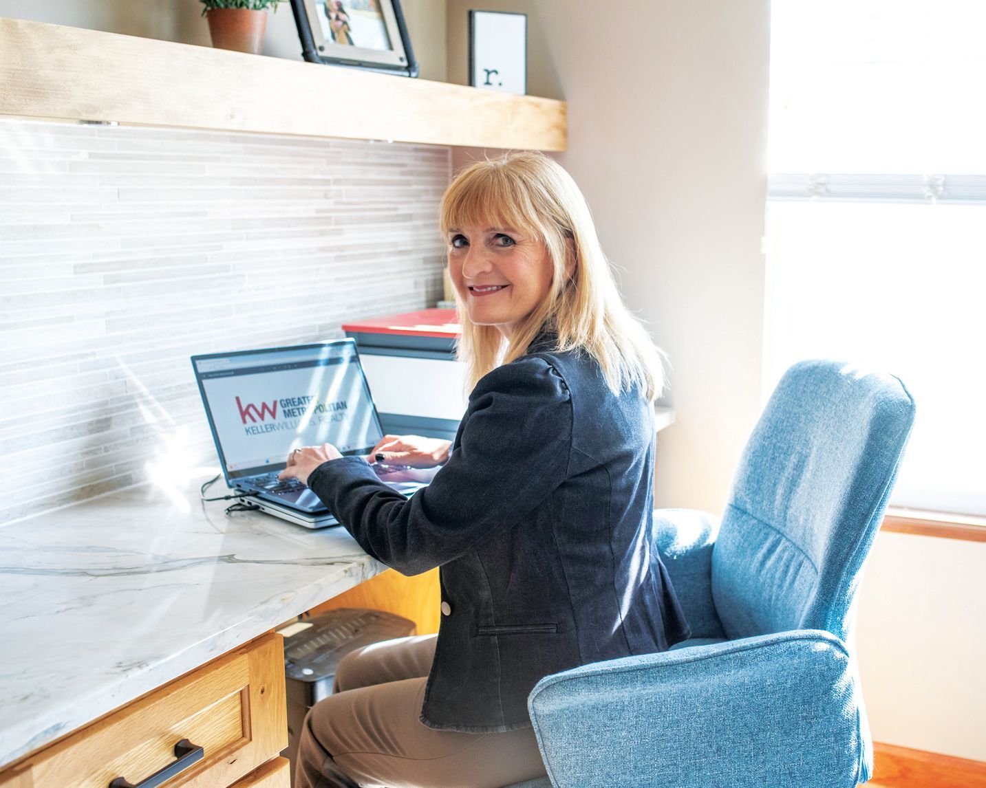 A professional seated at a home desk, smiling, typing on a laptop with a branded logo, in a bright, modern room.