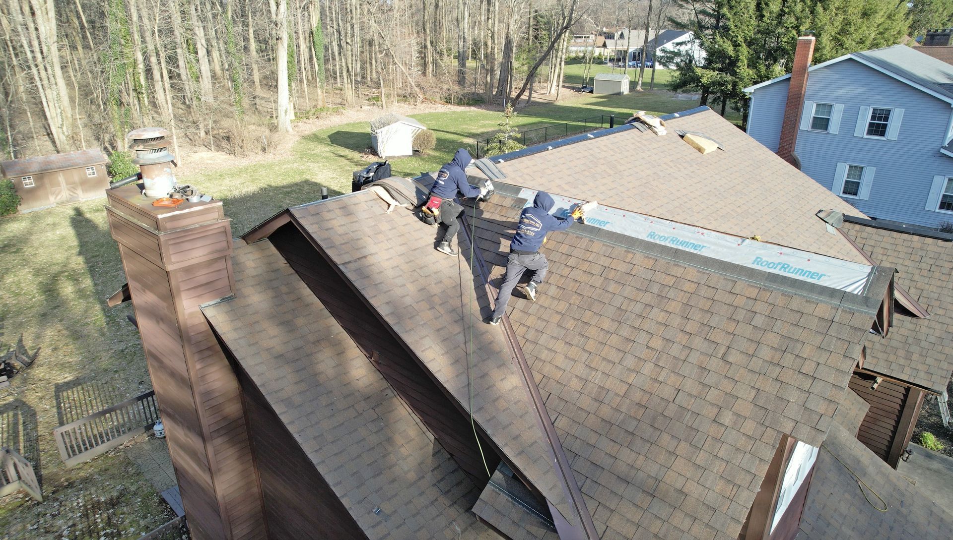 Two workers in blue uniforms install light-colored underlayment on a brown-shingled residential roof near a brick chimney.