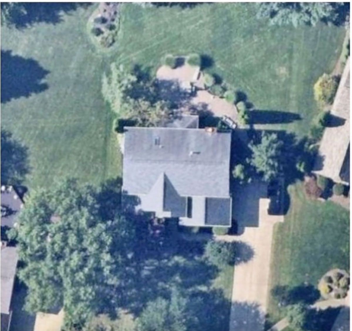 Aerial view of a residential house with a shingled roof, driveway, and surrounding lawn with trees.