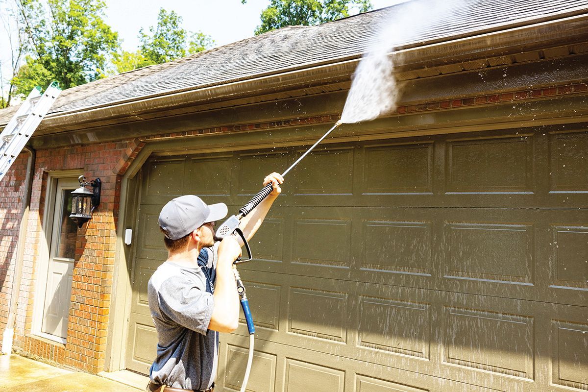 A person using a pressure washer to clean the exterior roof and garage door of a brick building.