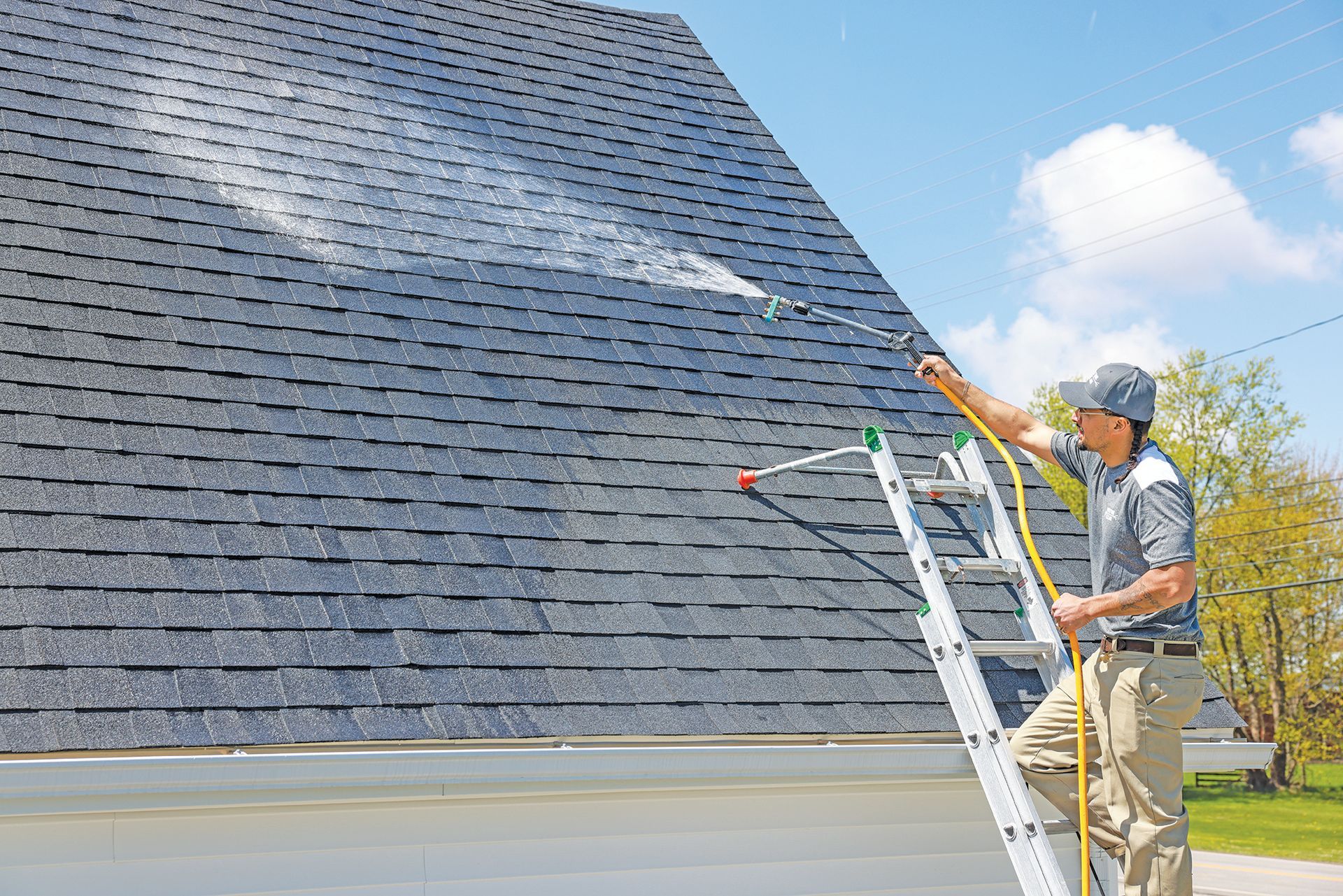 A worker in a gray shirt and tan pants stands on a ladder, spraying a dark shingled roof with water.