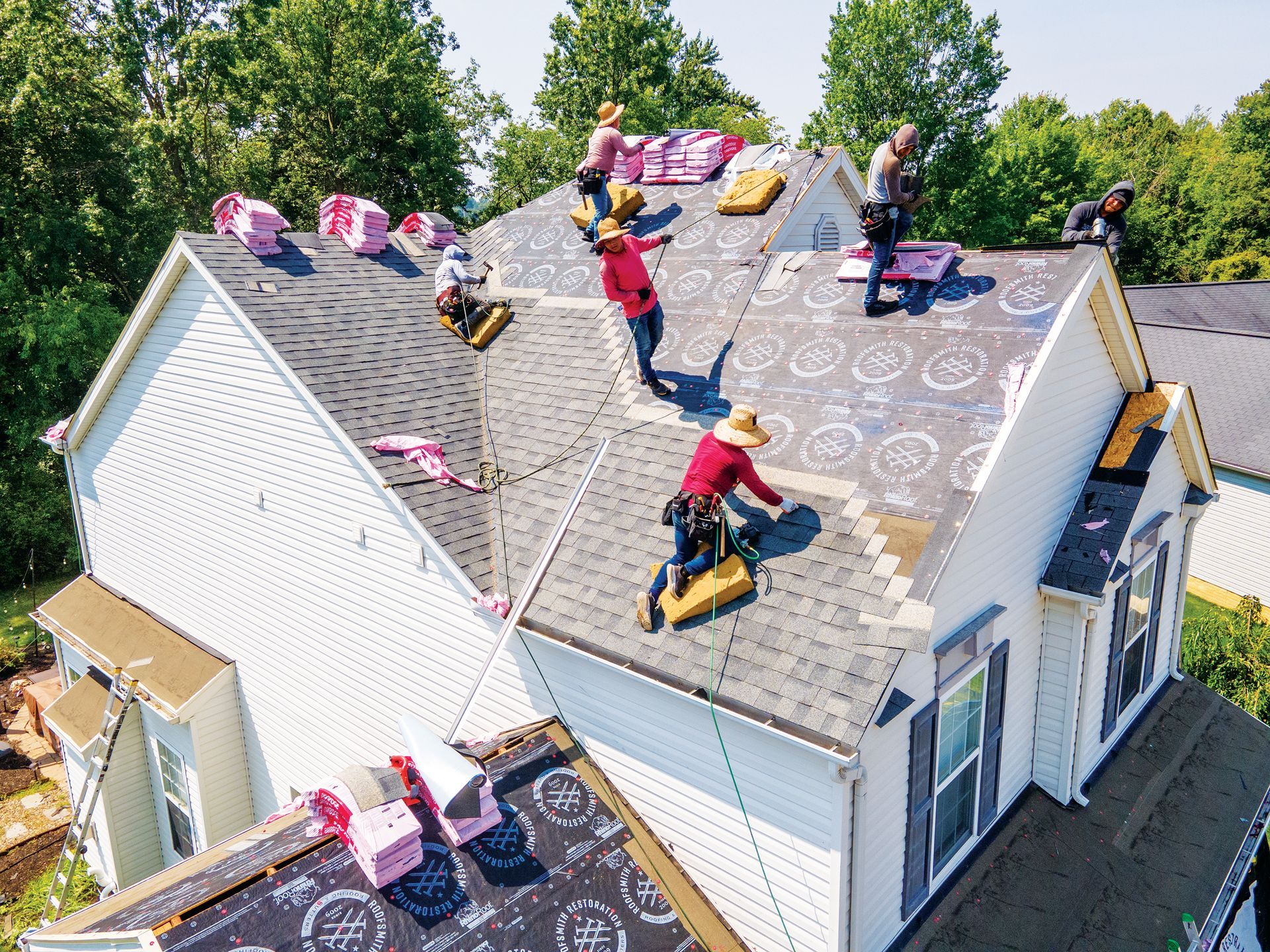 A crew of workers installing asphalt shingles and underlayment on the roof of a suburban home on a sunny day.