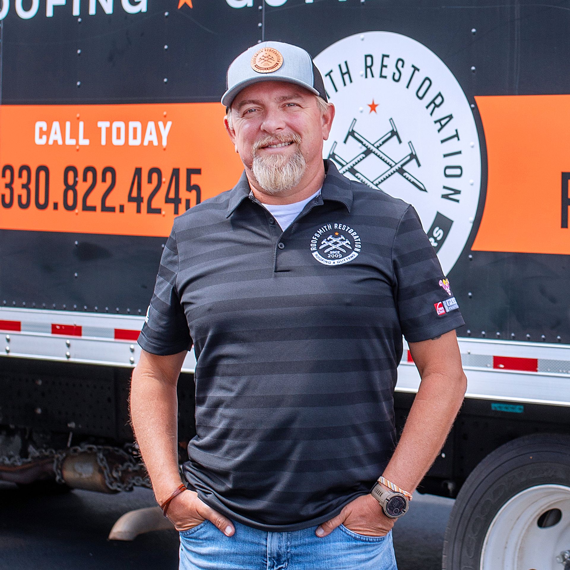 A man with a goatee wearing a gray cap and branded polo shirt stands in front of a company truck with contact information.