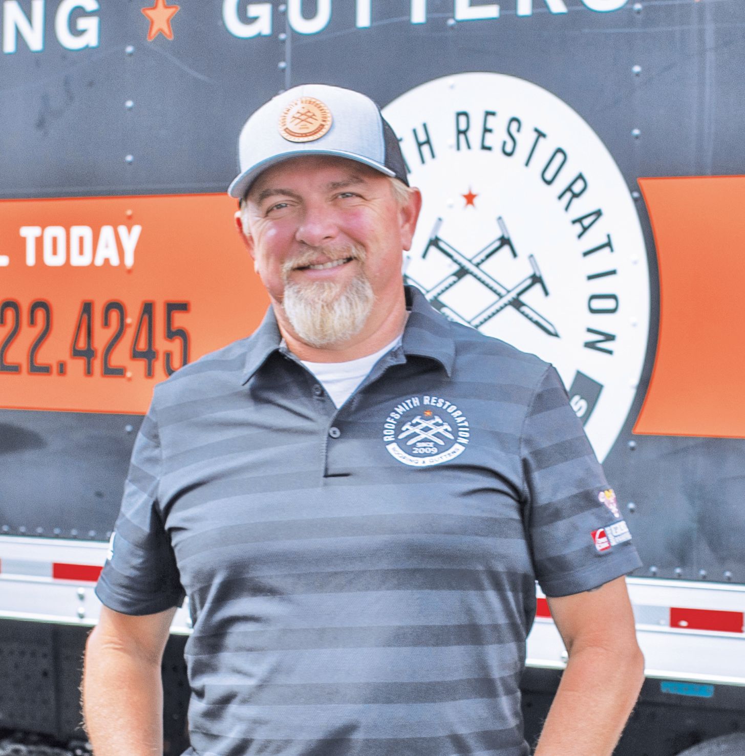 A smiling person wearing a branded cap and polo shirt, standing in front of a company truck with roofing logos.