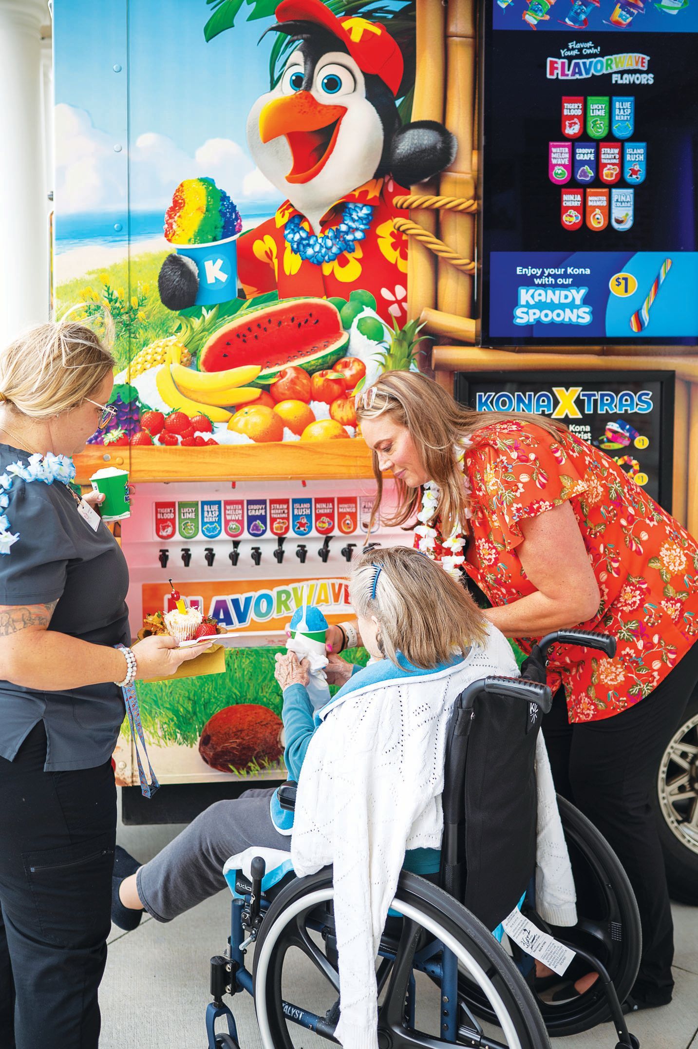 People interact at a colorful shaved ice stand featuring a cartoon mascot, fruit illustrations, and flavor taps.