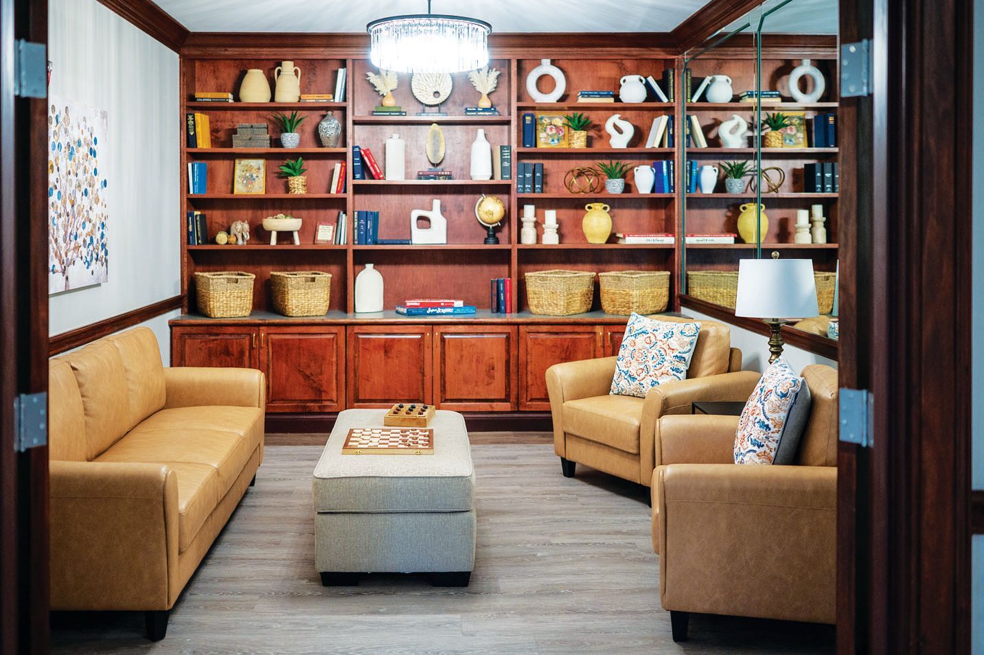 A cozy lounge featuring tan leather seating, an ottoman with a game board, and large wooden shelves filled with decor.