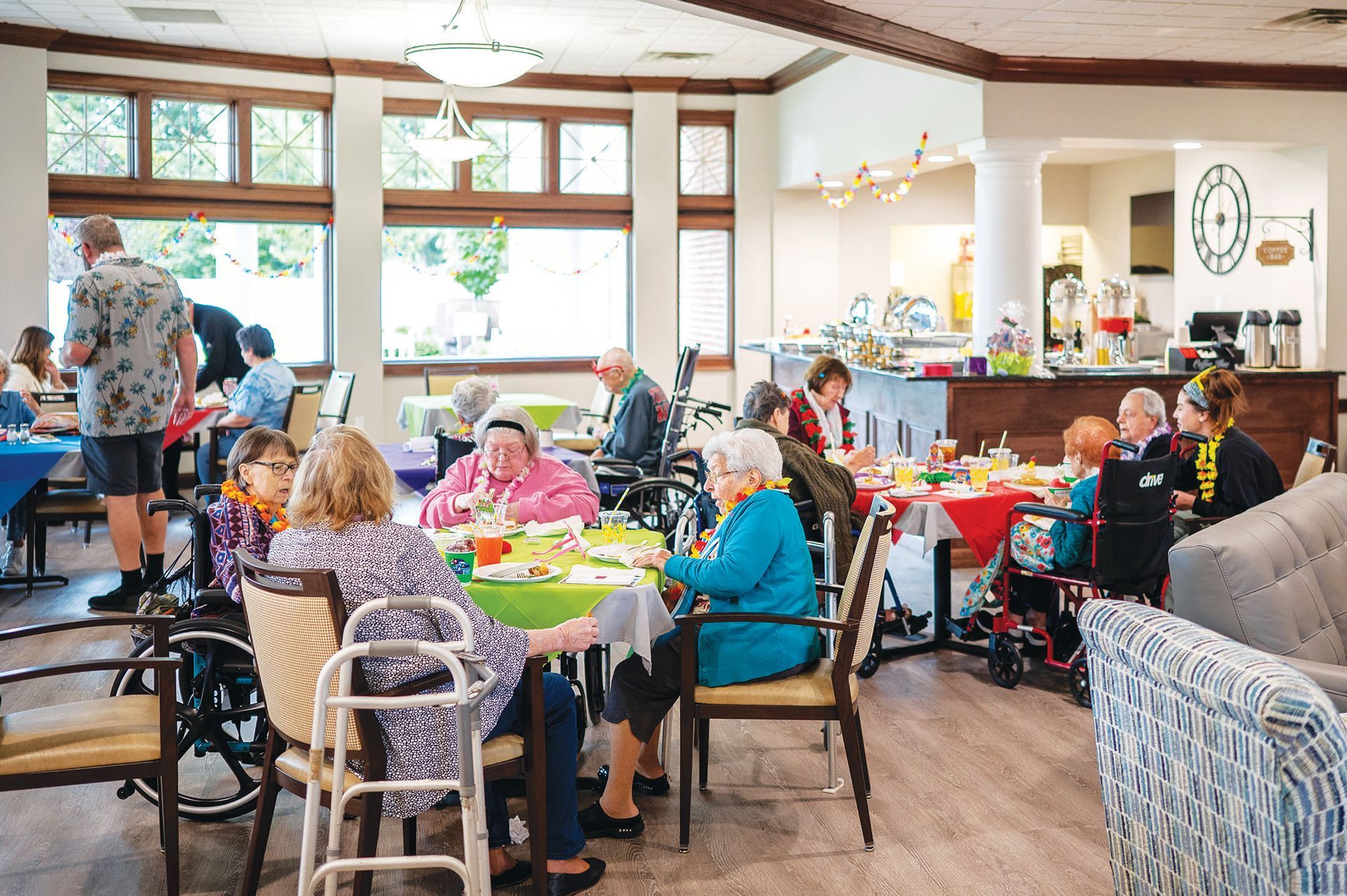 People dine and socialize in a bright, sunlit dining room with large windows, tables, and a service counter in the back.