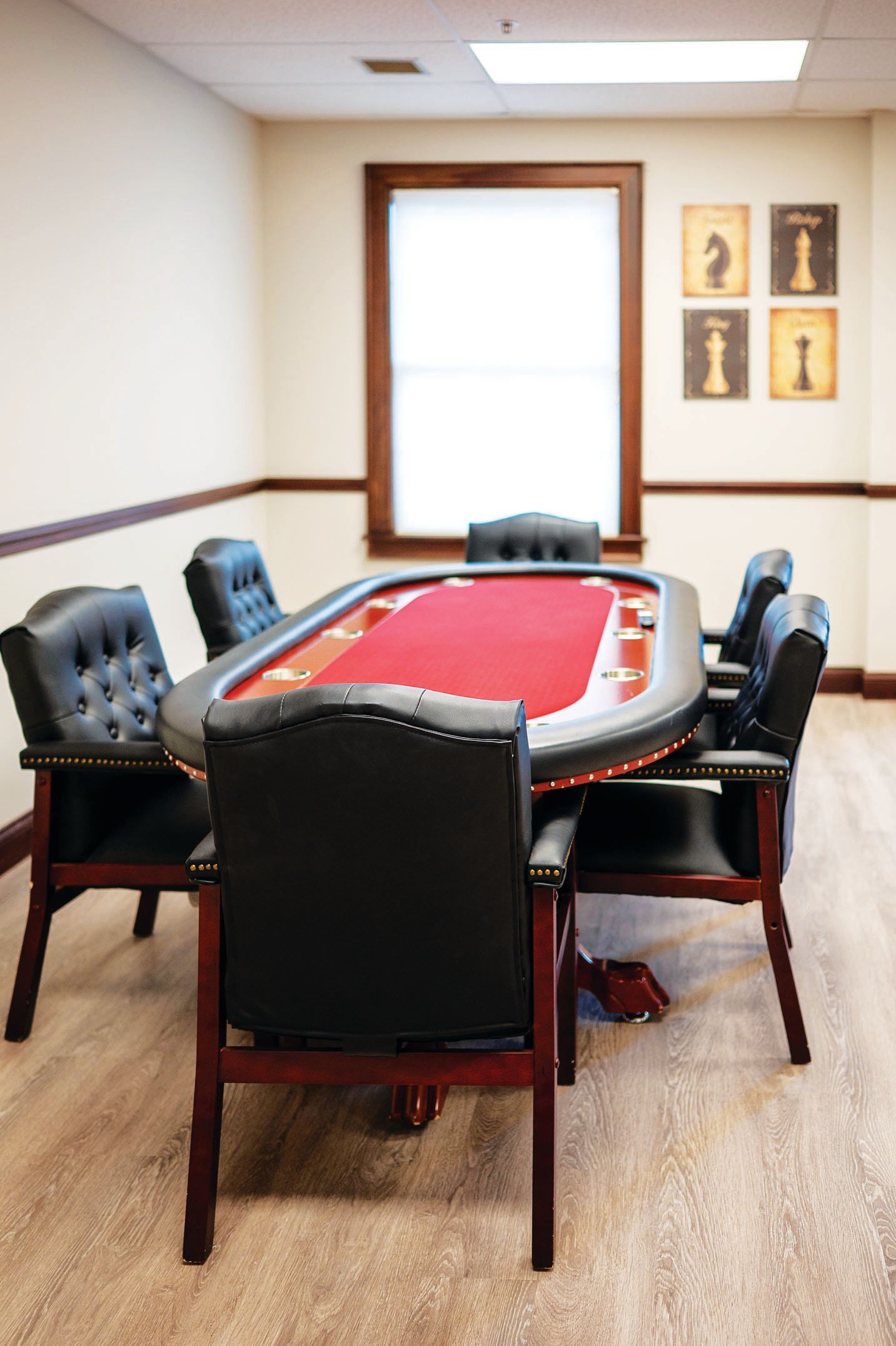 A poker table with a red felt top and black chairs in a room with a window and four chess piece wall prints.